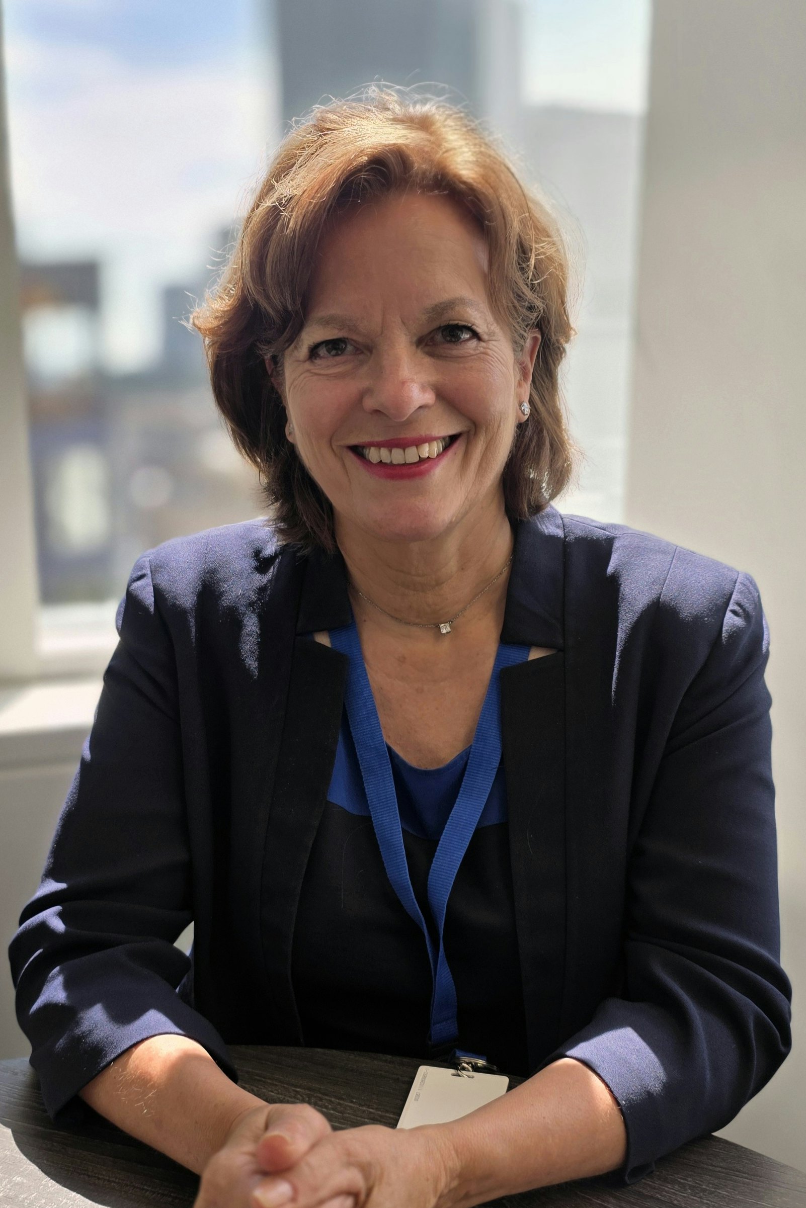 Portrait professionnel d’une femme souriante, assise près d’une fenêtre. Elle porte une veste bleu foncé et un lanyard bleu, avec la lumière naturelle éclairant son visage.