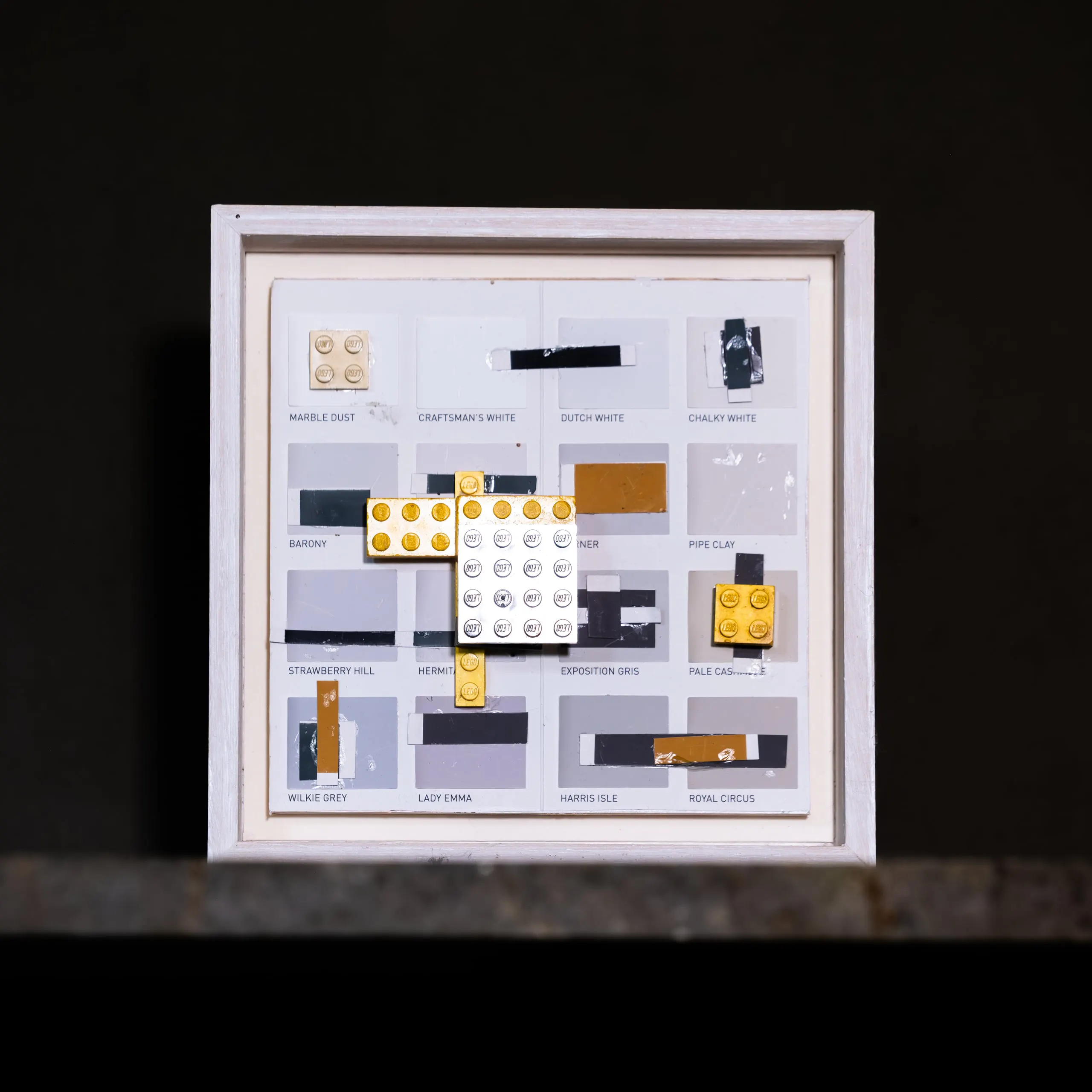 A small square framed work of grey swatches with white and yellow LEGO across the centre