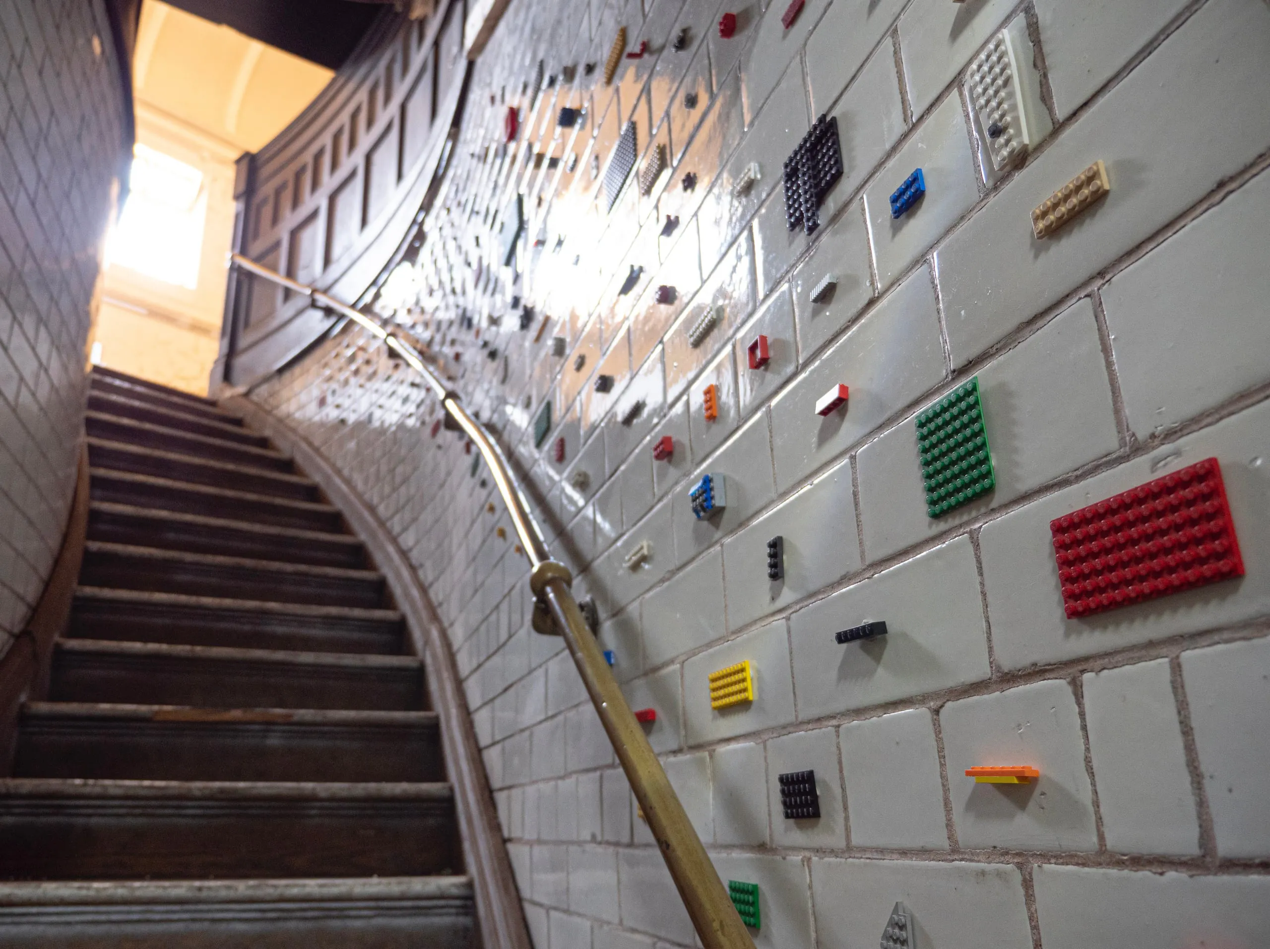 A curving stairwell of white glazed tile in the former magistrates’ court, coloured LEGO plates set one to a tile beside a brass handrail