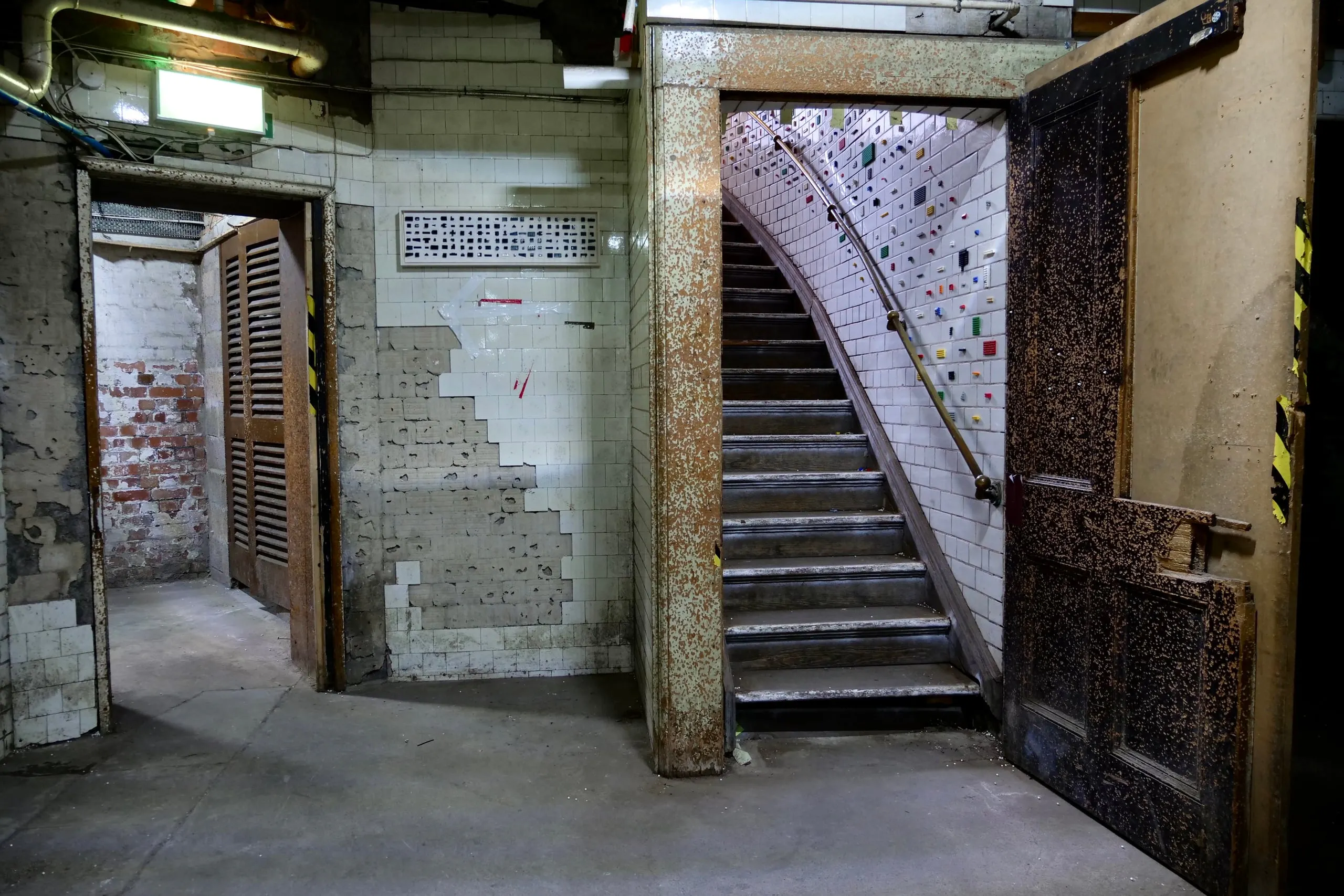 A hall in the former magistrates’ court: a doorway to bare brick, a stripped tile wall hung with a framed chart work, and a tiled stair set with coloured LEGO