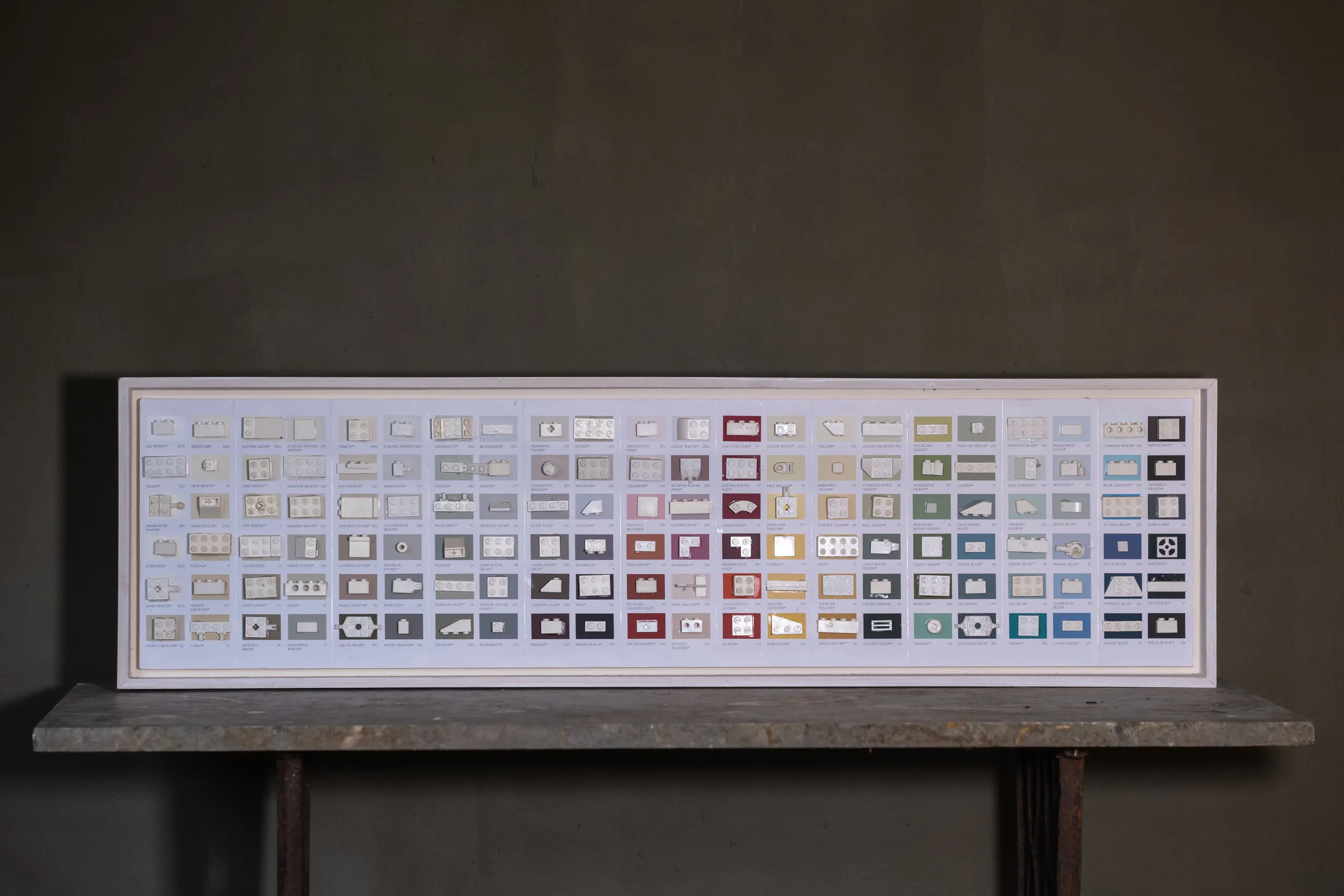 A long framed panel of paint-chart segments, each carrying a small LEGO piece, resting on a trestle table