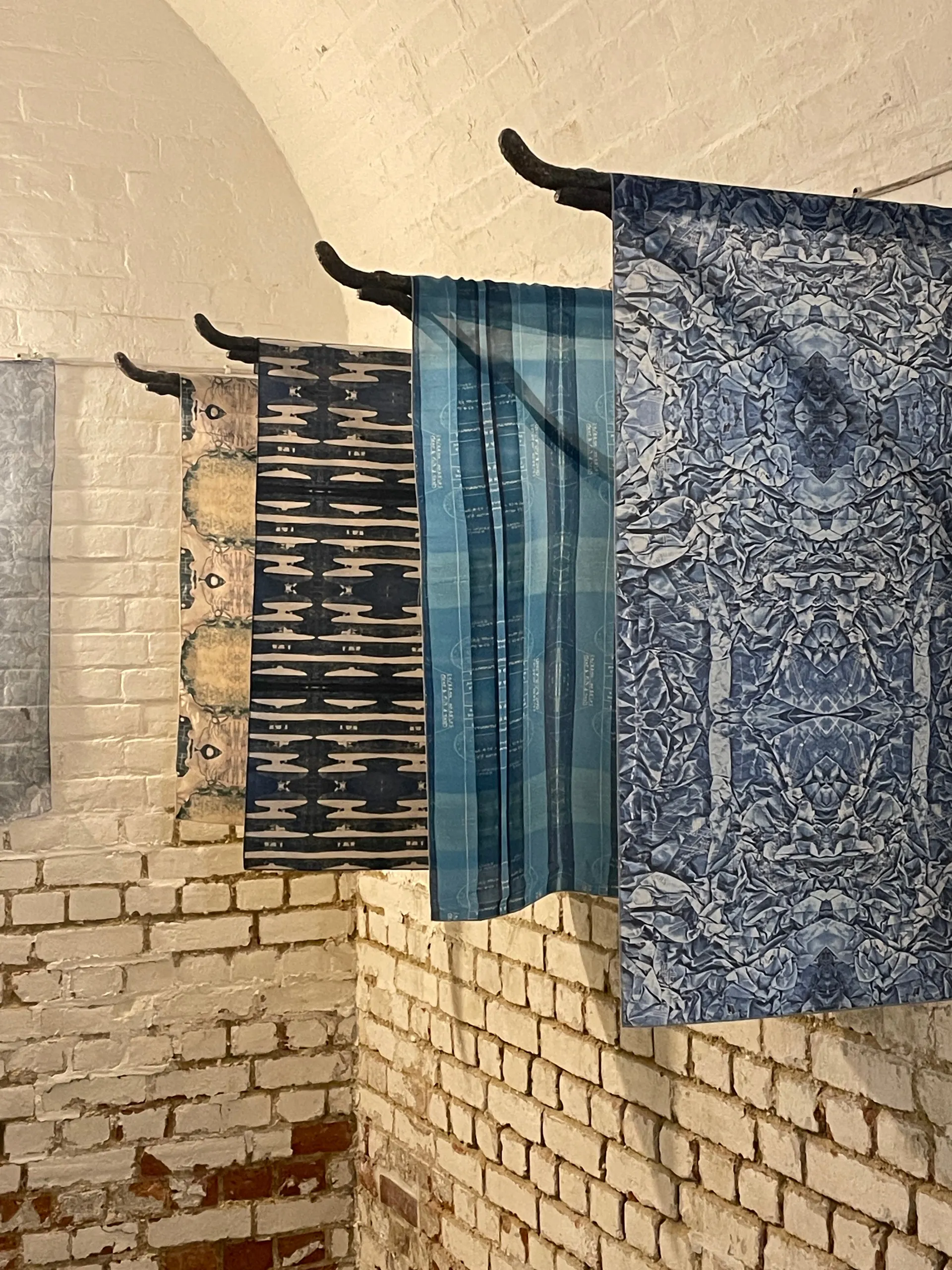 Four printed textile panels hung on iron brackets in a whitewashed brick vault
