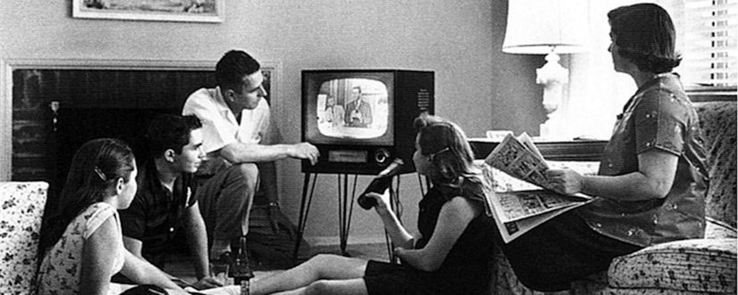 Photo of a family gathered around an old black and white television set. 