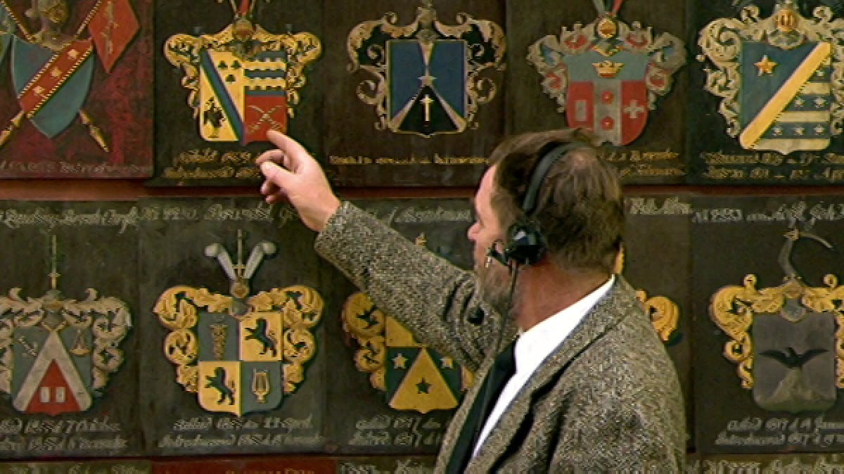 TV presenter pointing at old-fashioned crest 