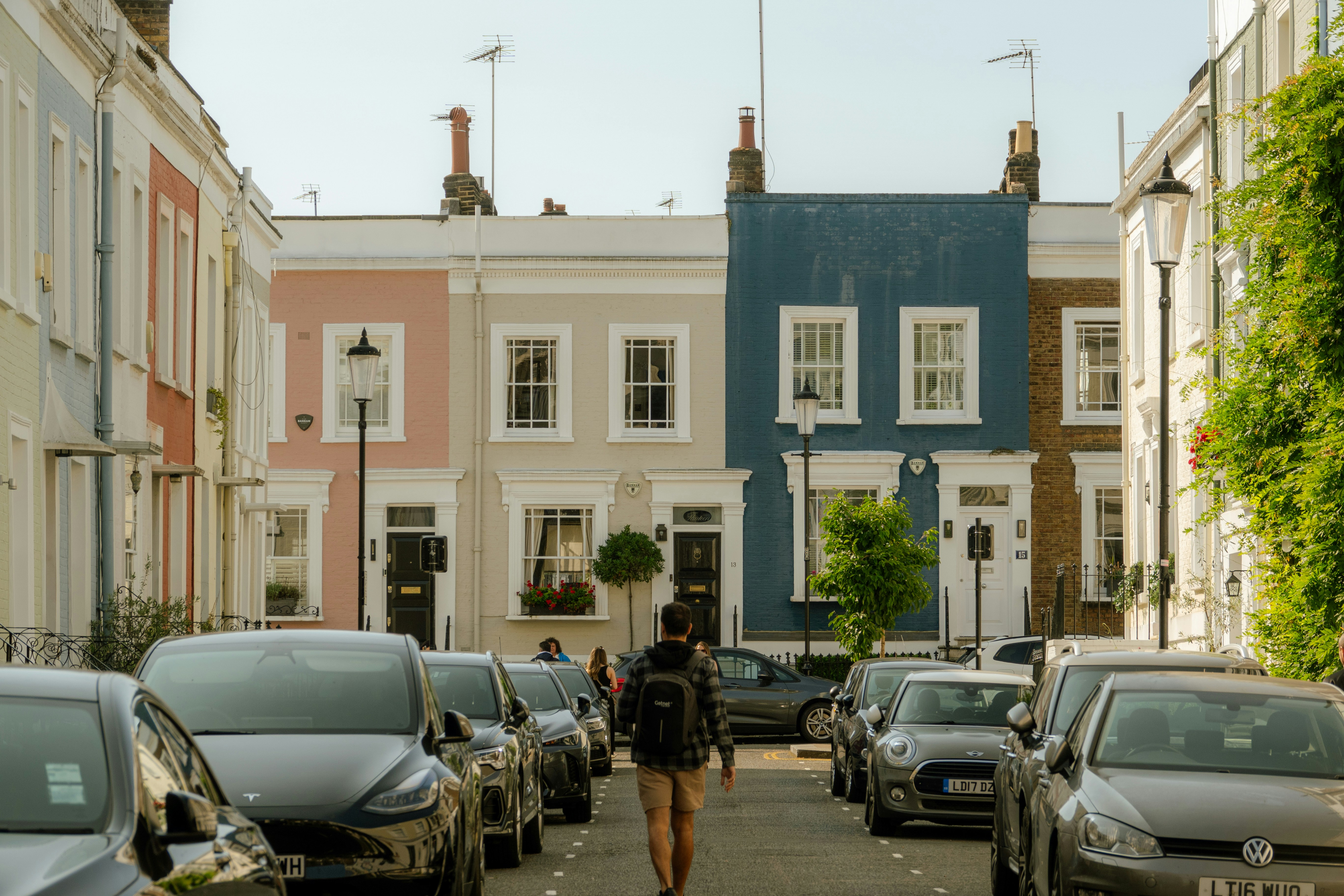 A quiet UK residential street with pastel-coloured terraced houses, parked cars on both sides, and a person walking away from the camera.