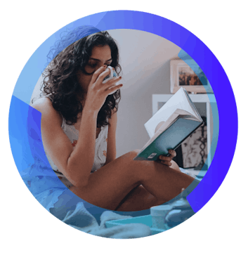 mulher com cabelos cacheados de pijama, sentada da cama lendo um livro e tomando café