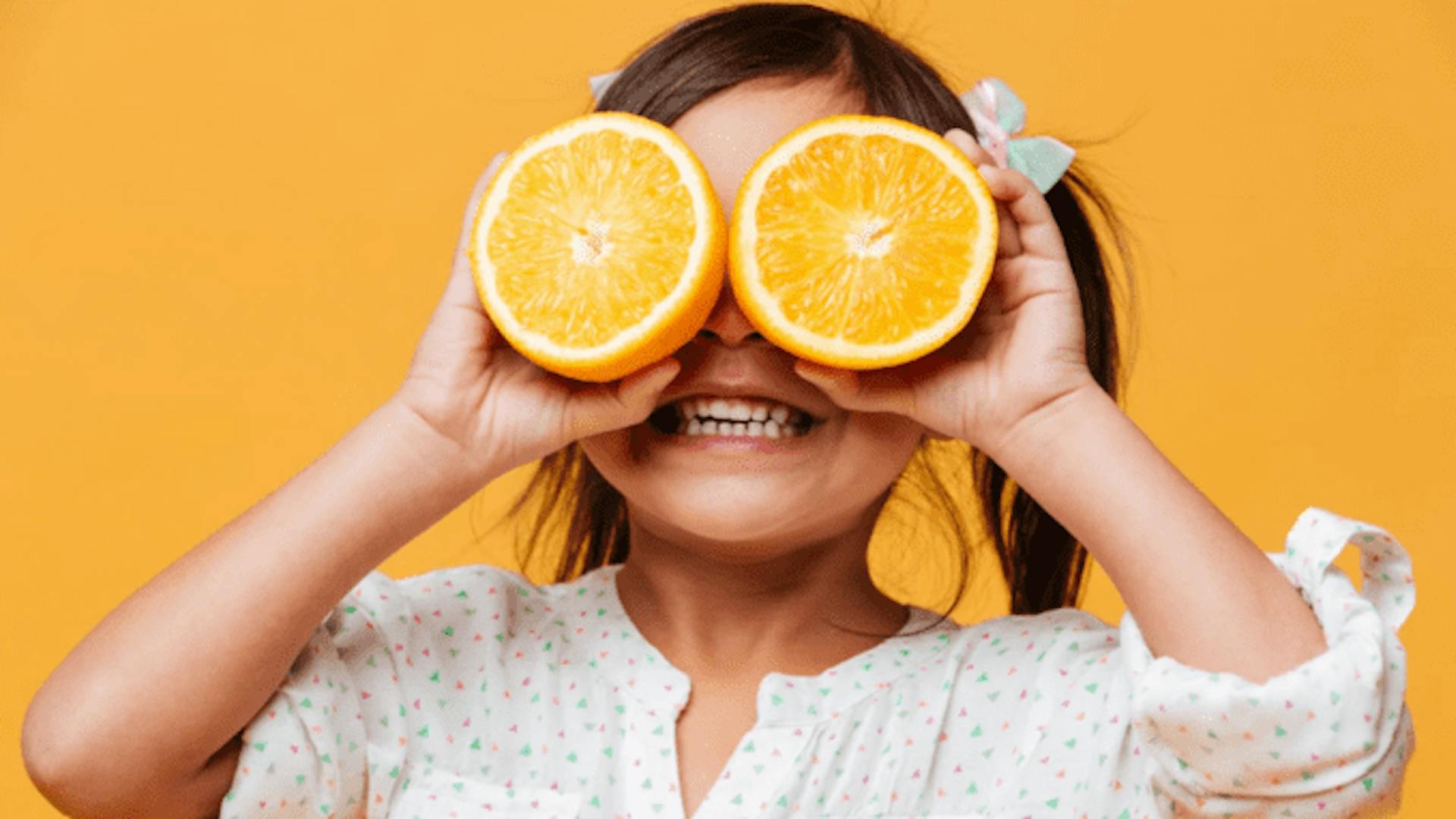 criança pequena sorridente, cobrindo os olhos com laranjas e parede na cor laranja ao fundo