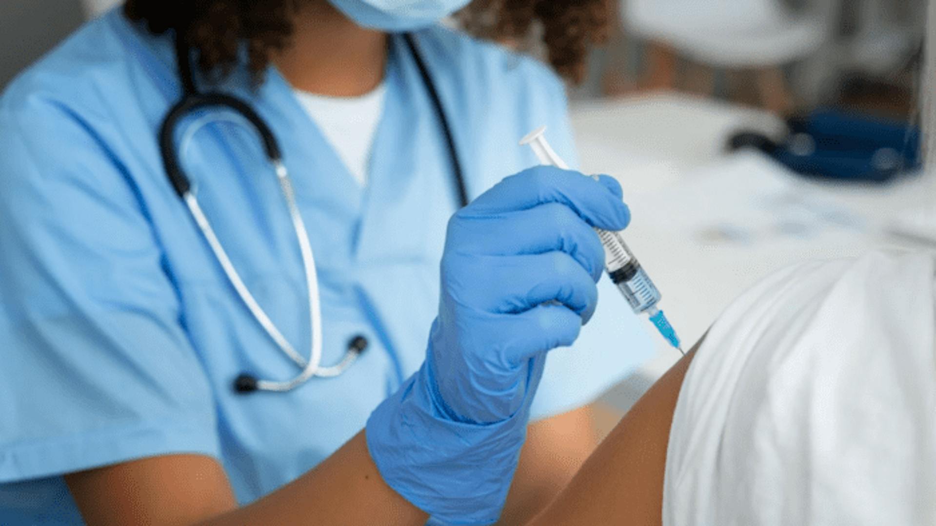 Enfermeira vestida com roupa, máscara e luvas azuis, aplica vacina em braço de paciente