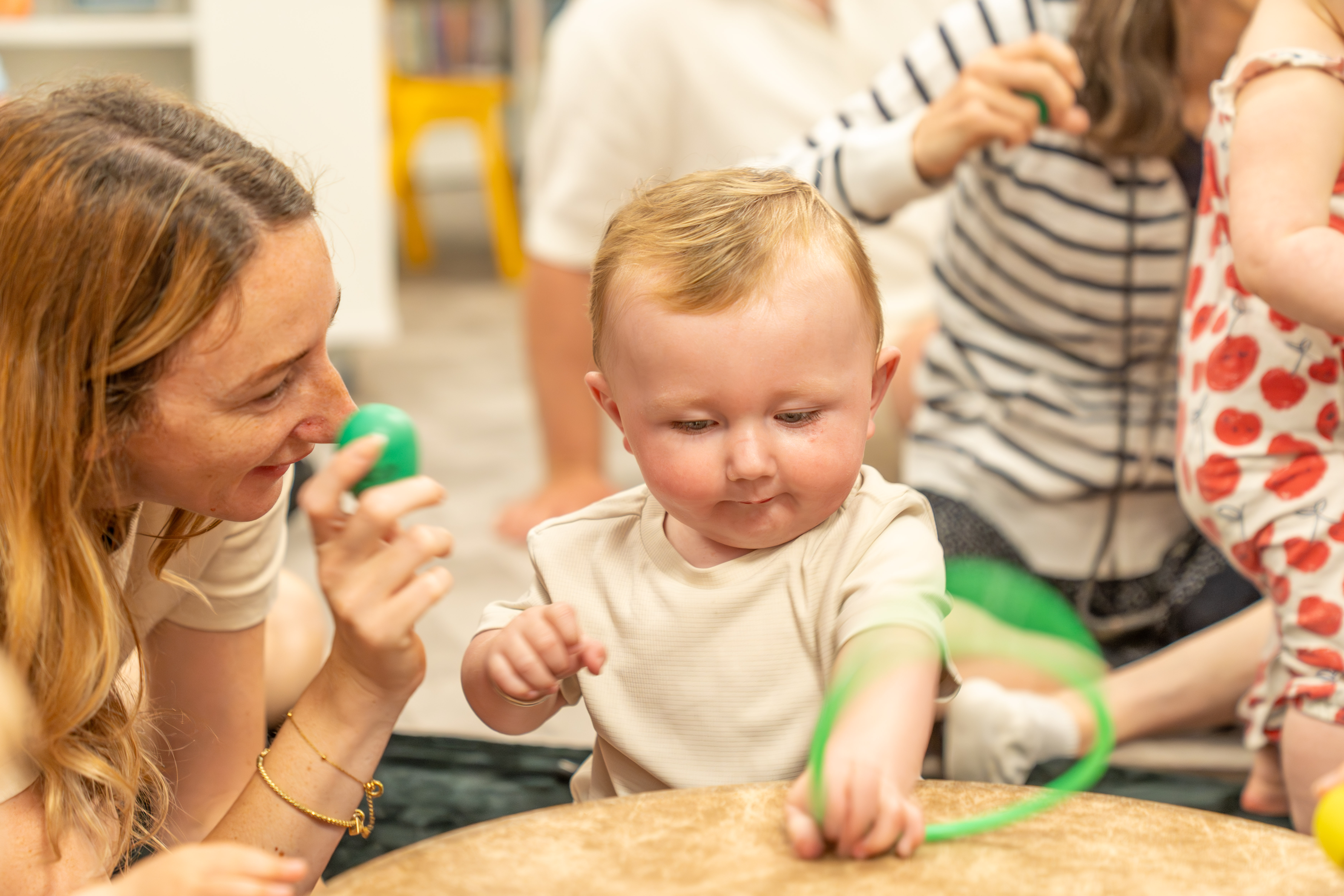 A mother and baby engaging. She is holding a green egg shaped shaker and the baby is holding green material with their hand on a drum