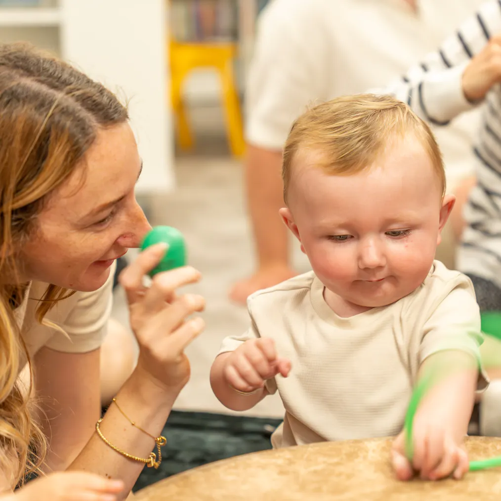 A mother and baby engaging. She is holding a green egg shaped shaker and the baby is holding green material with their hand on a drum