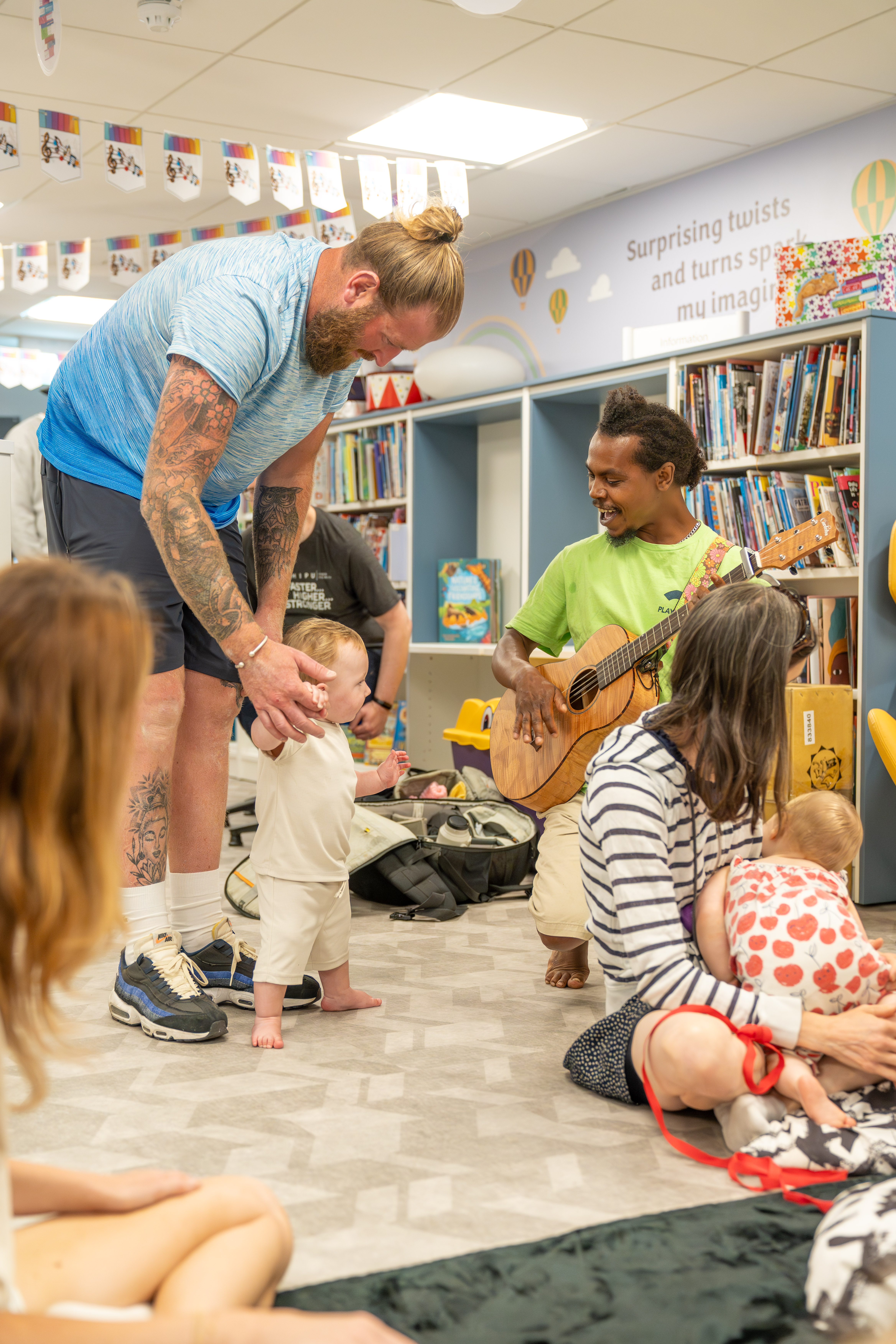 Babies, young children, parents/carers and an artist on a mat during a Family Playground session. They are in a library, surrounded by book shelves. 
