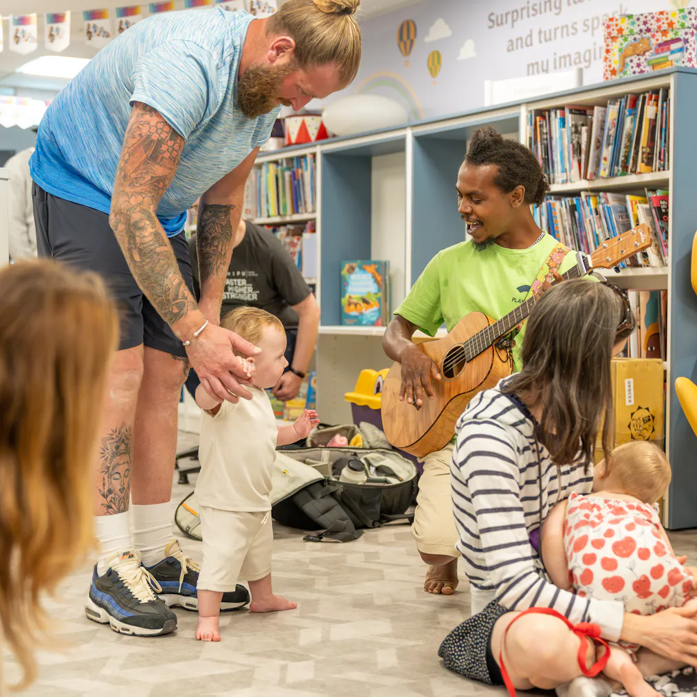Babies, young children, parents/carers and an artist on a mat during a Family Playground session. They are in a library, surrounded by book shelves.