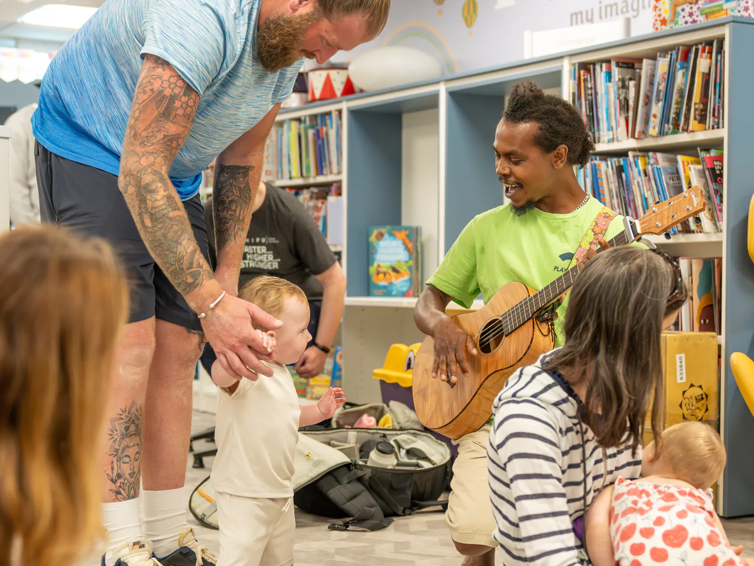 Babies, young children, parents/carers and an artist on a mat during a Family Playground session. They are in a library, surrounded by book shelves.