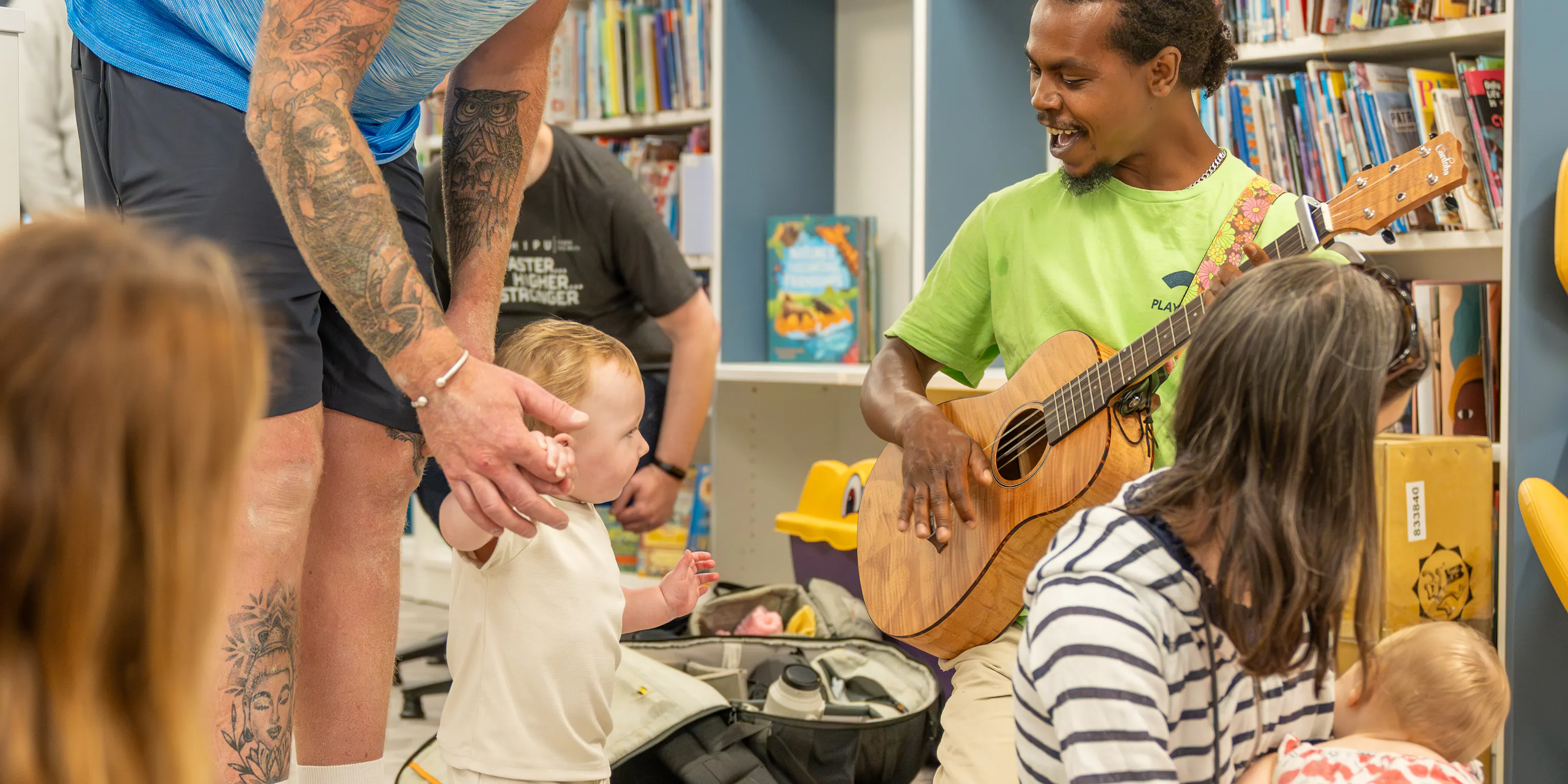 Babies, young children, parents/carers and an artist on a mat during a Family Playground session. They are in a library, surrounded by book shelves.