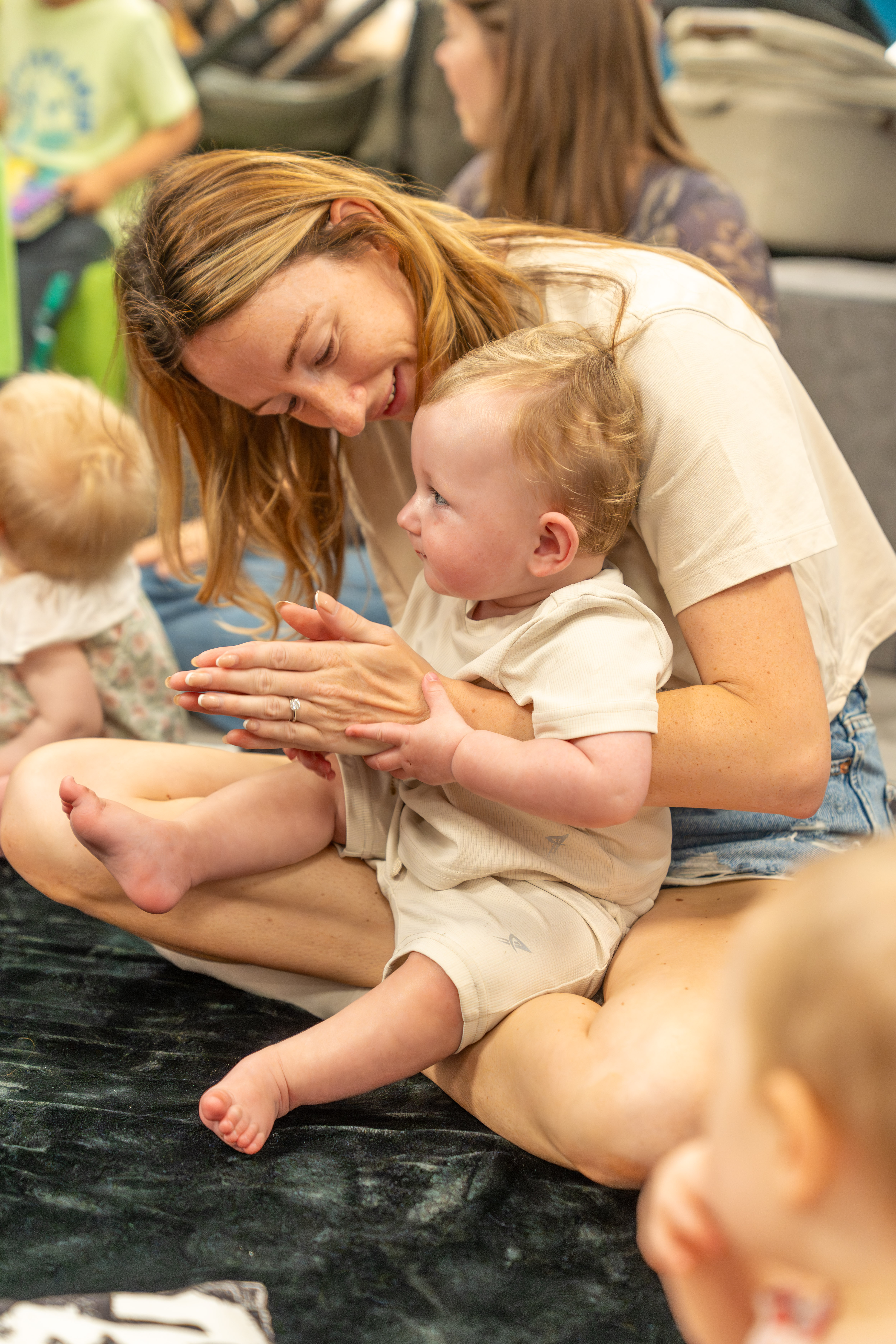 A mother and baby sat together on a black mat. They are surrounded by other babies and parents/carers.