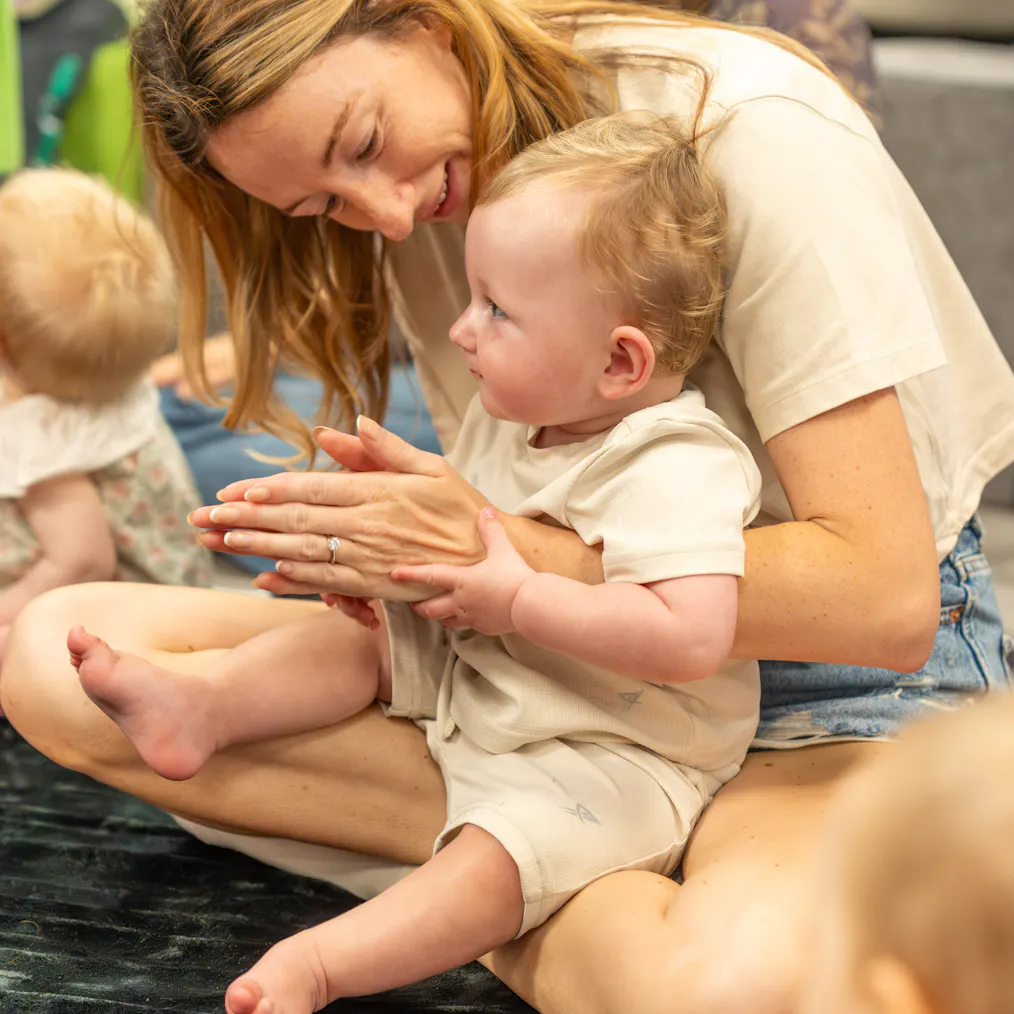 A mother and baby sat together on a black mat. They are surrounded by other babies and parents/carers.