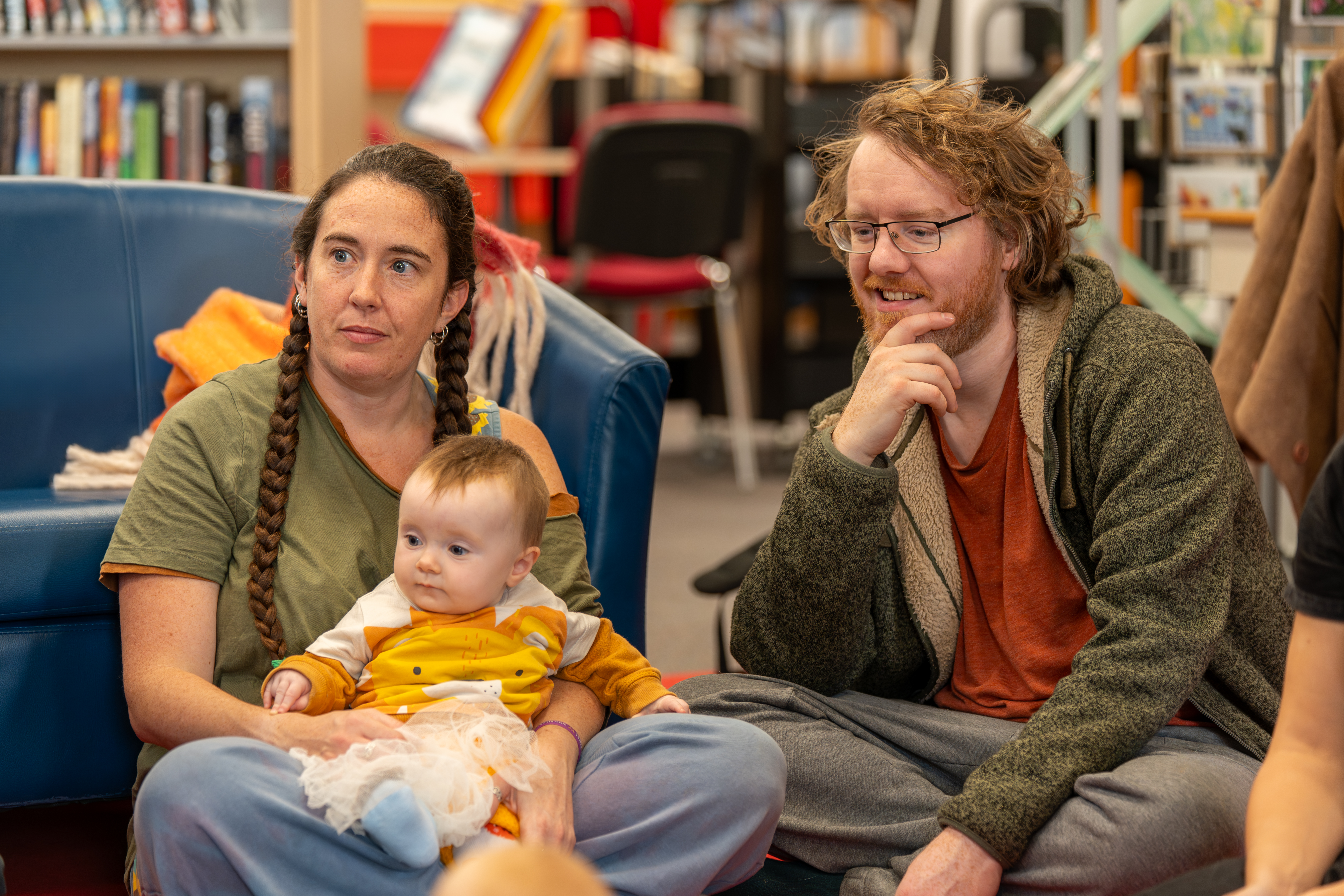 Two parents/carers and a baby sat together and looking in the same direction 
