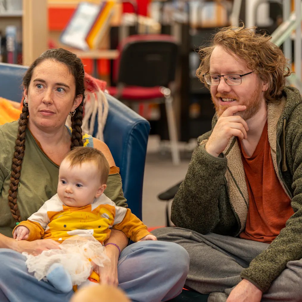 Two parents/carers and a baby sat together and looking in the same direction