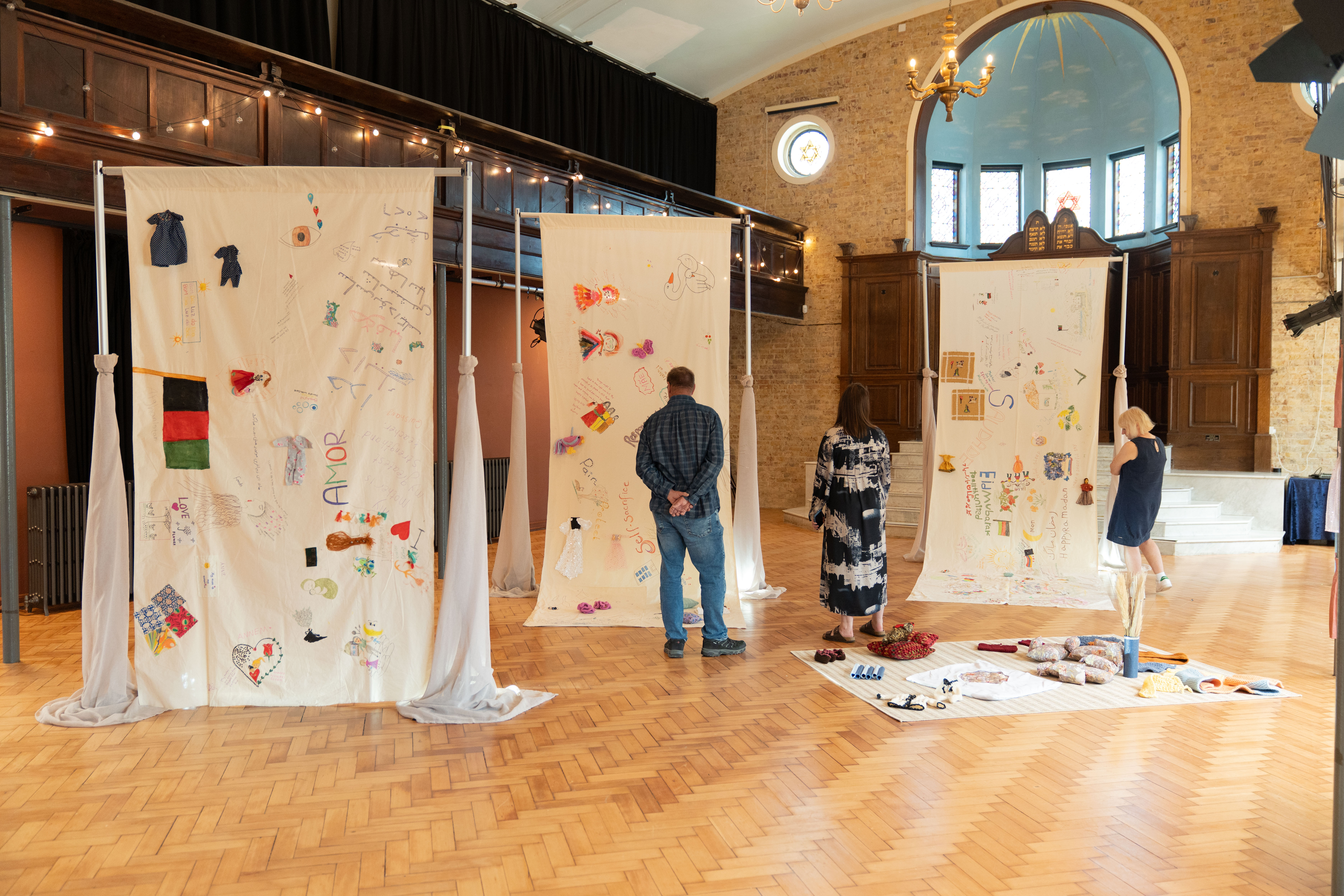 People stranding in an exhibition space. There are tall pieces of clothes hanging with paintings and illustrations