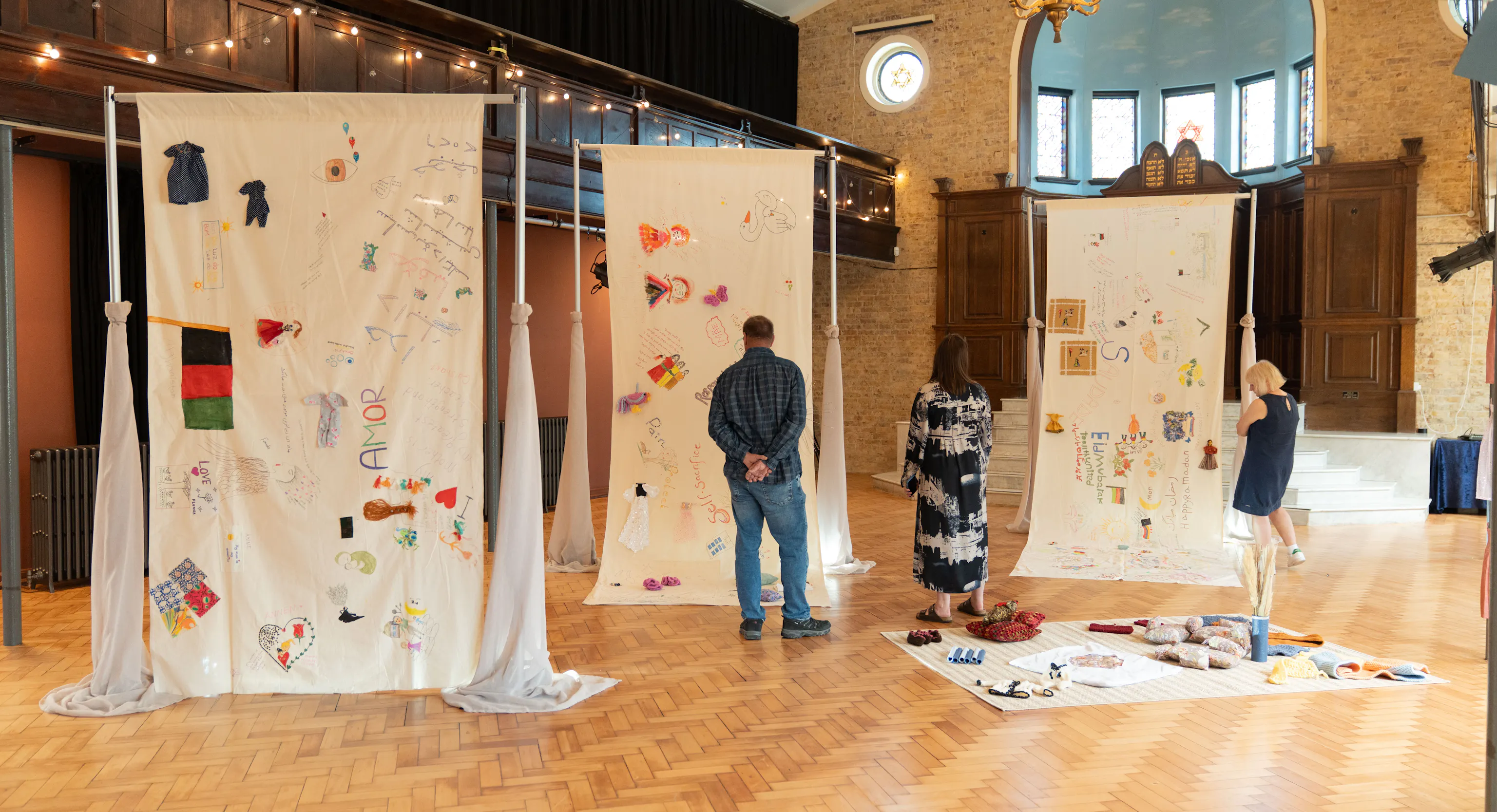 People stranding in an exhibition space. There are tall pieces of clothes hanging with paintings and illustrations