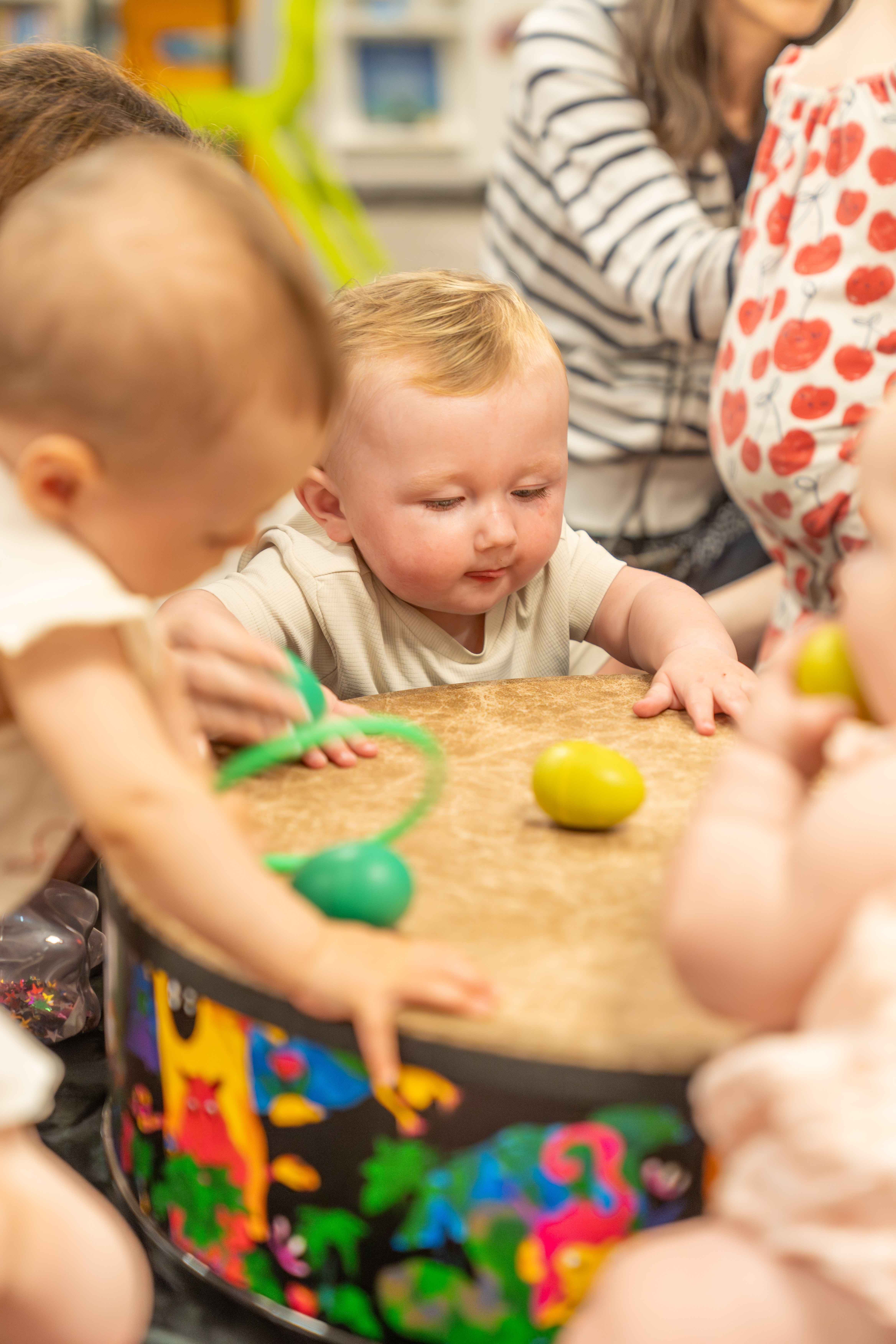A group of babies and young children gather in front of a colourful drum with egg shaped rattles on it 