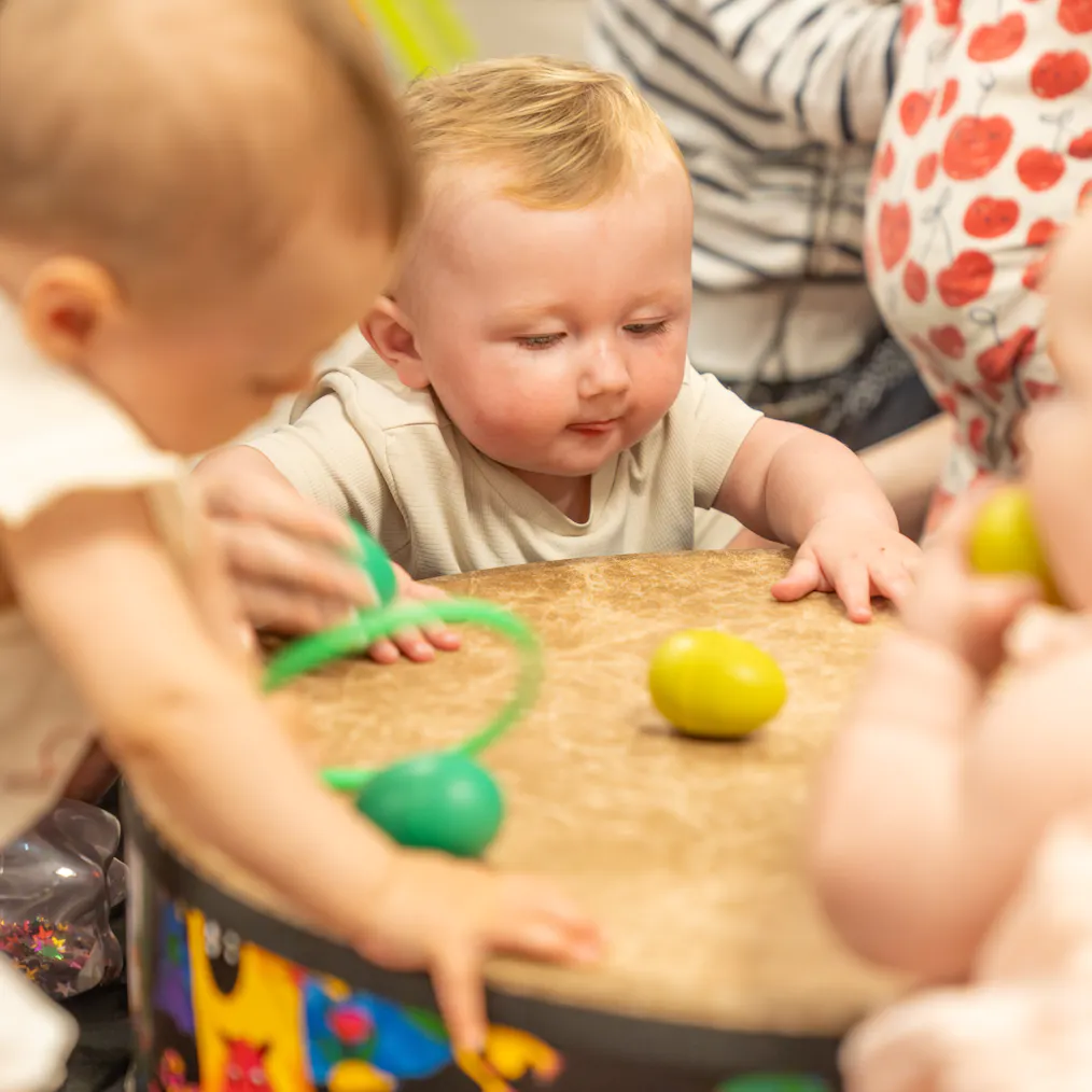 A group of babies and young children gather in front of a colourful drum with egg shaped rattles on it