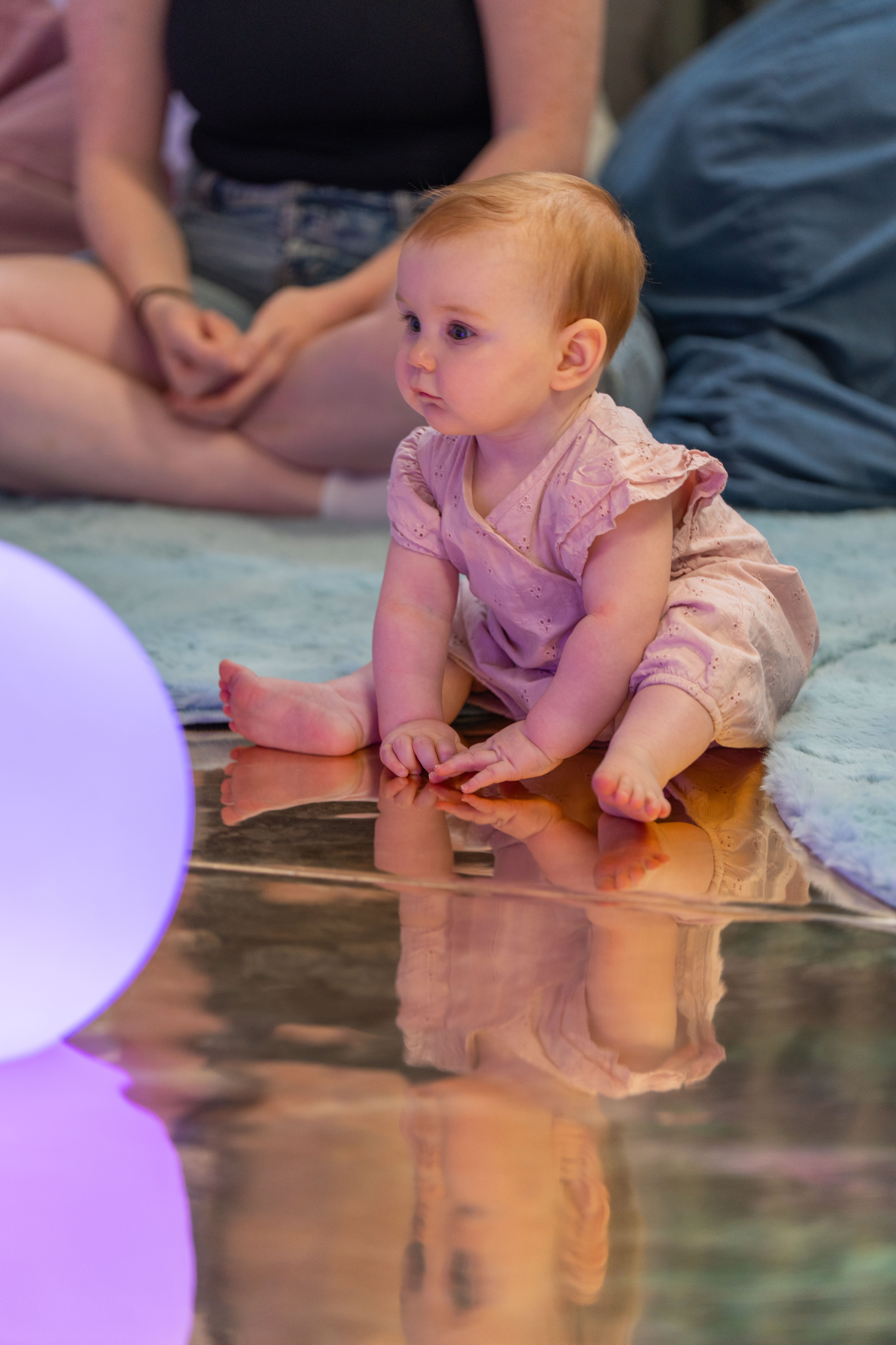 A baby sat on a reflective floor looking at a performance. Next to them is a big bright lilac ball 