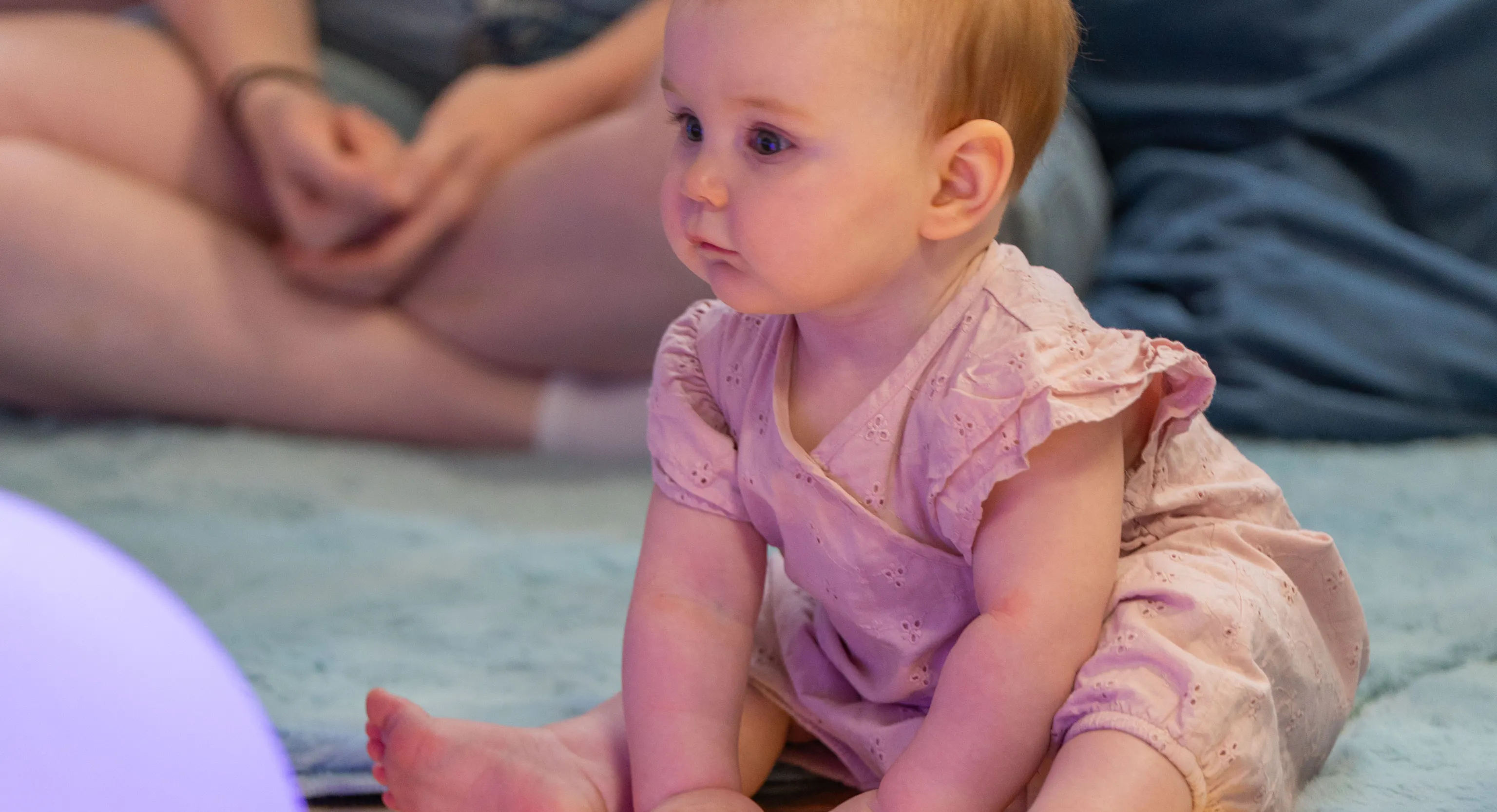 A baby sat on a reflective floor looking at a performance. Next to them is a big bright lilac ball