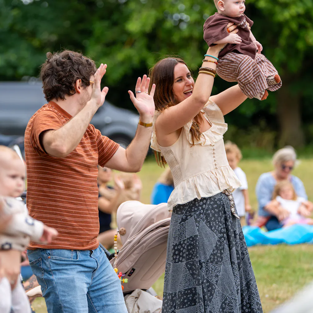 Two parents and a baby dancing to a song. One is holding up their hand and the other holding up the baby