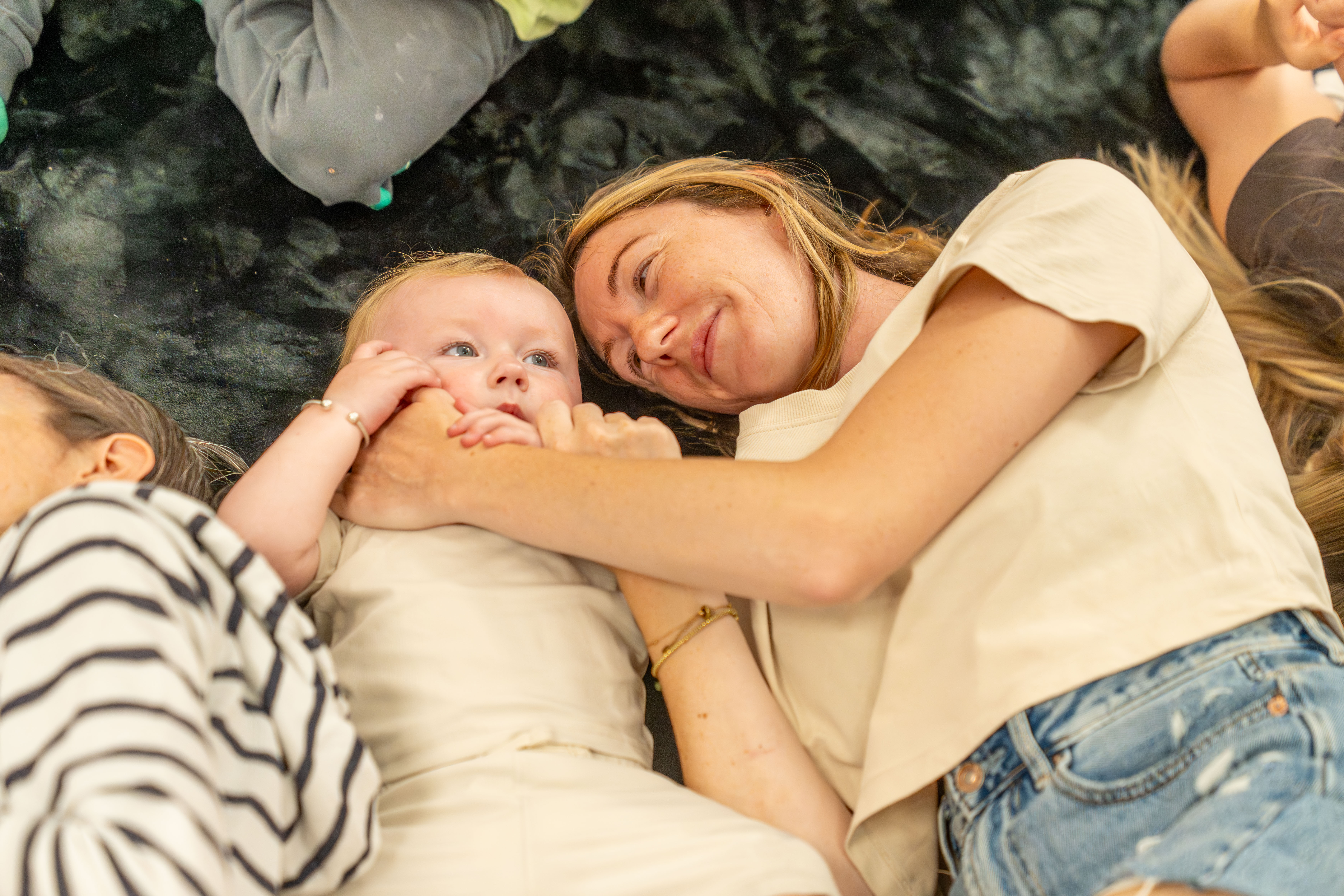 A young child and mother lying on a black mat. The parent/carer has their hand across the child's face and the child is touching their hand