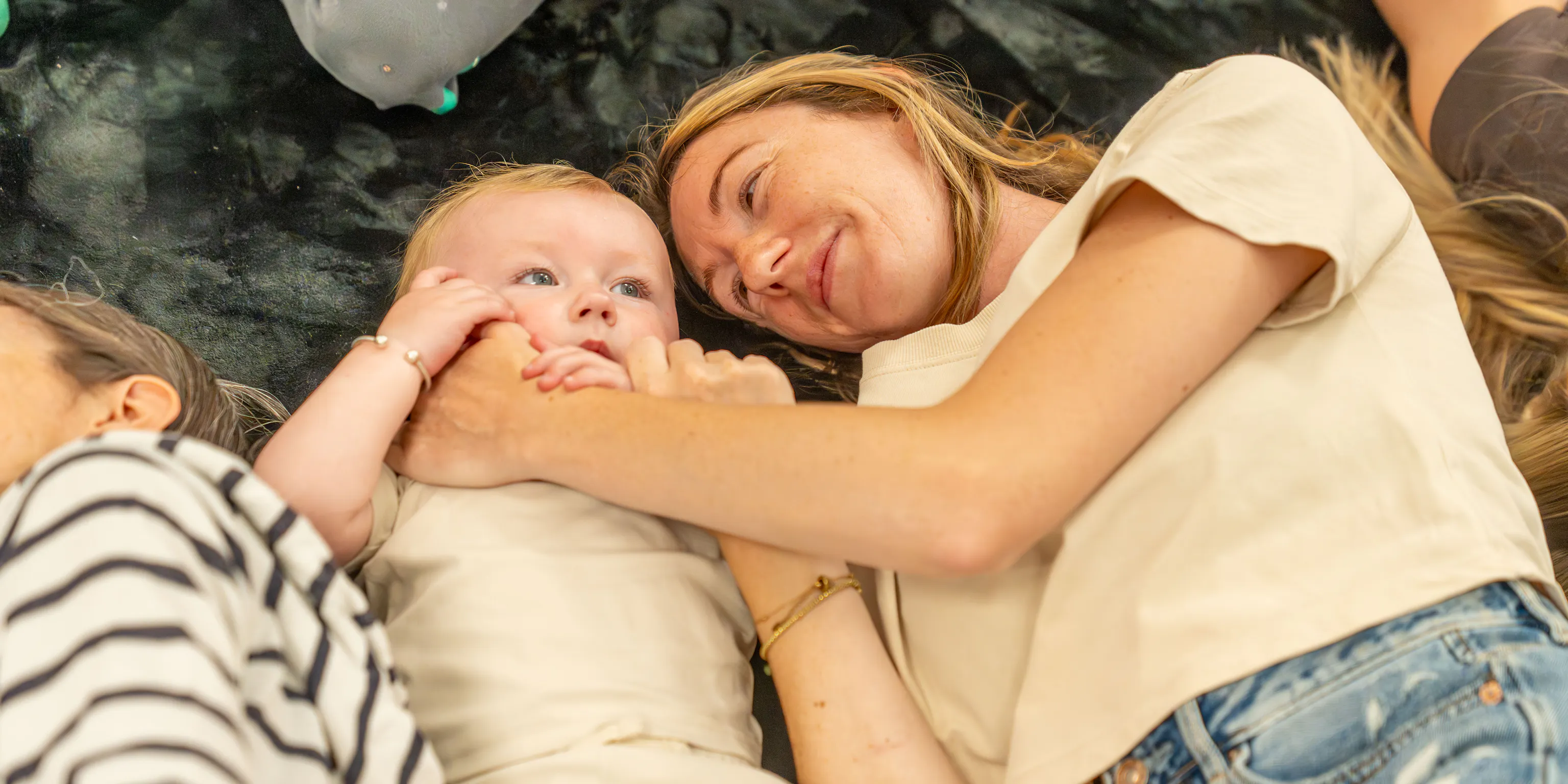 A young child and mother lying on a black mat. The parent/carer has their hand across the child's face and the child is touching their hand