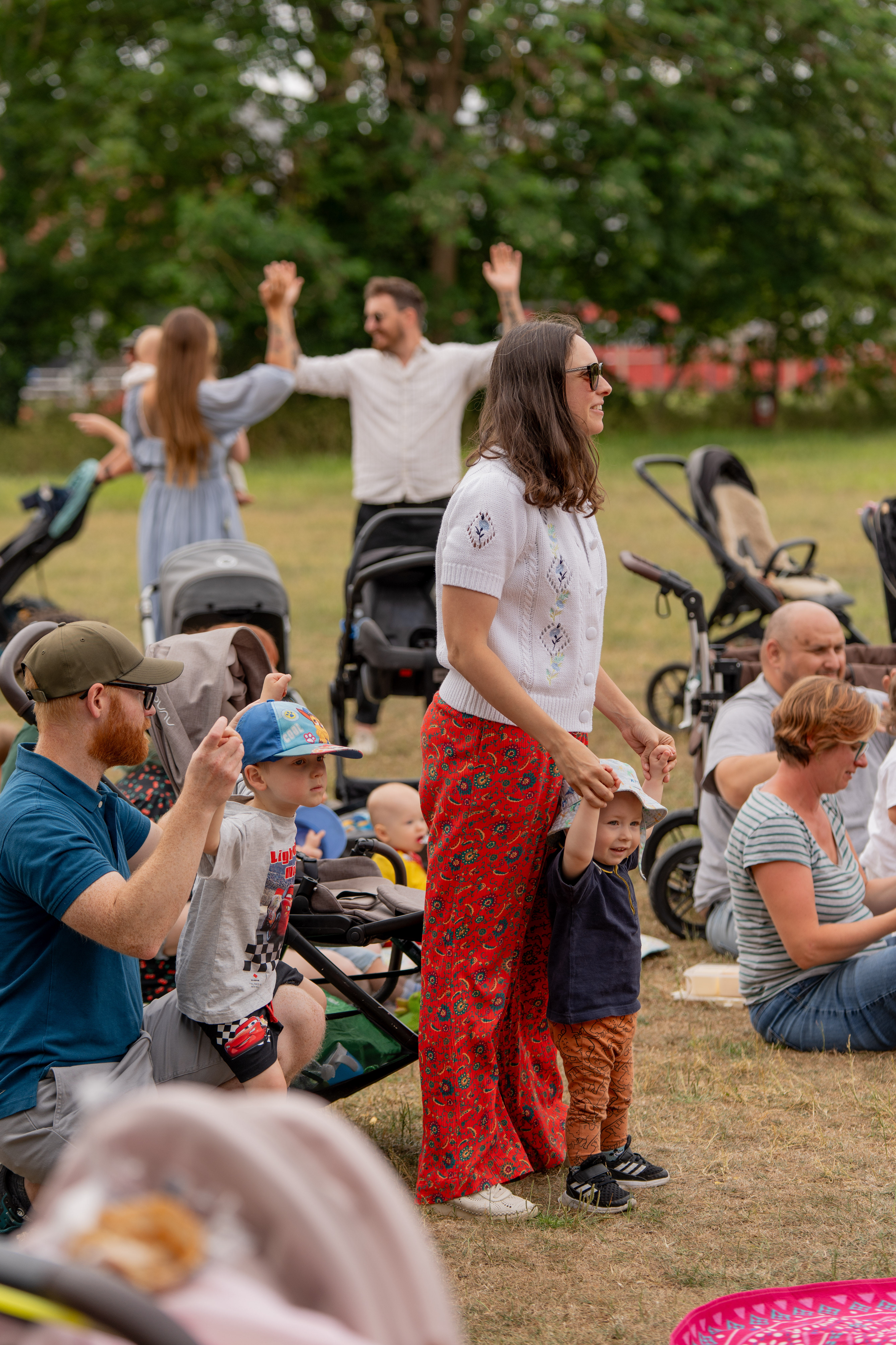 Families in an outdoor park. Some are standing, sitting and others have their hands up 