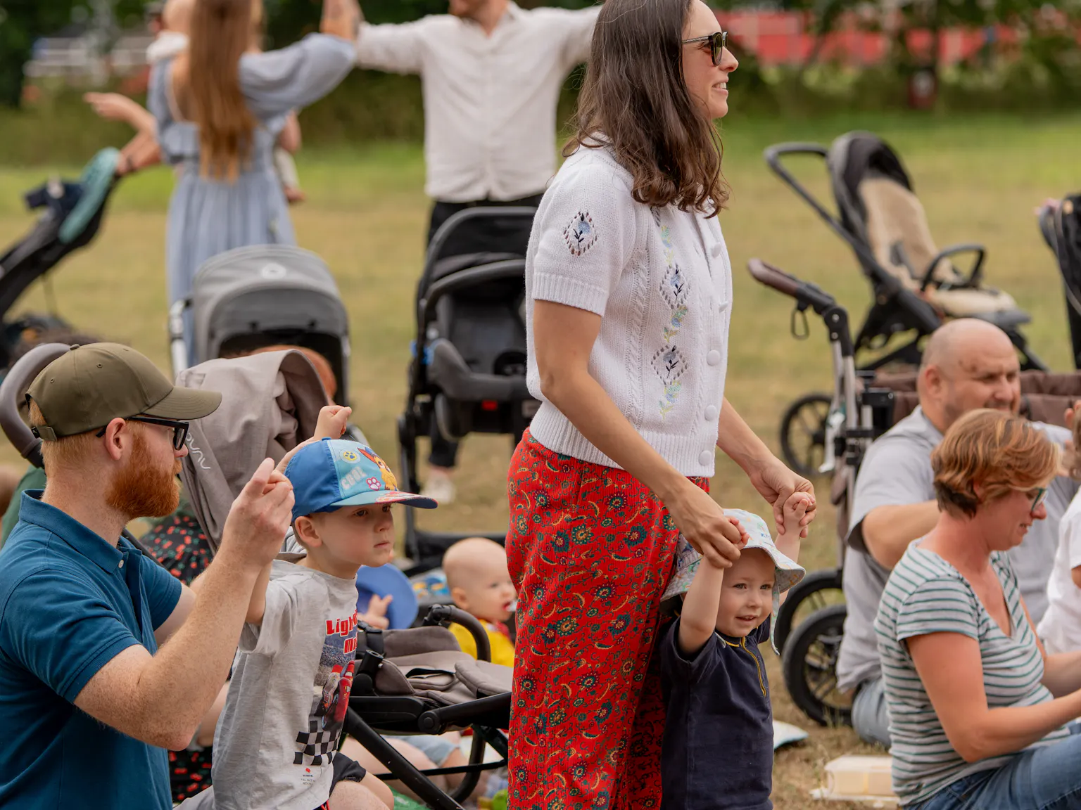 Families in an outdoor park. Some are standing, sitting and others have their hands up