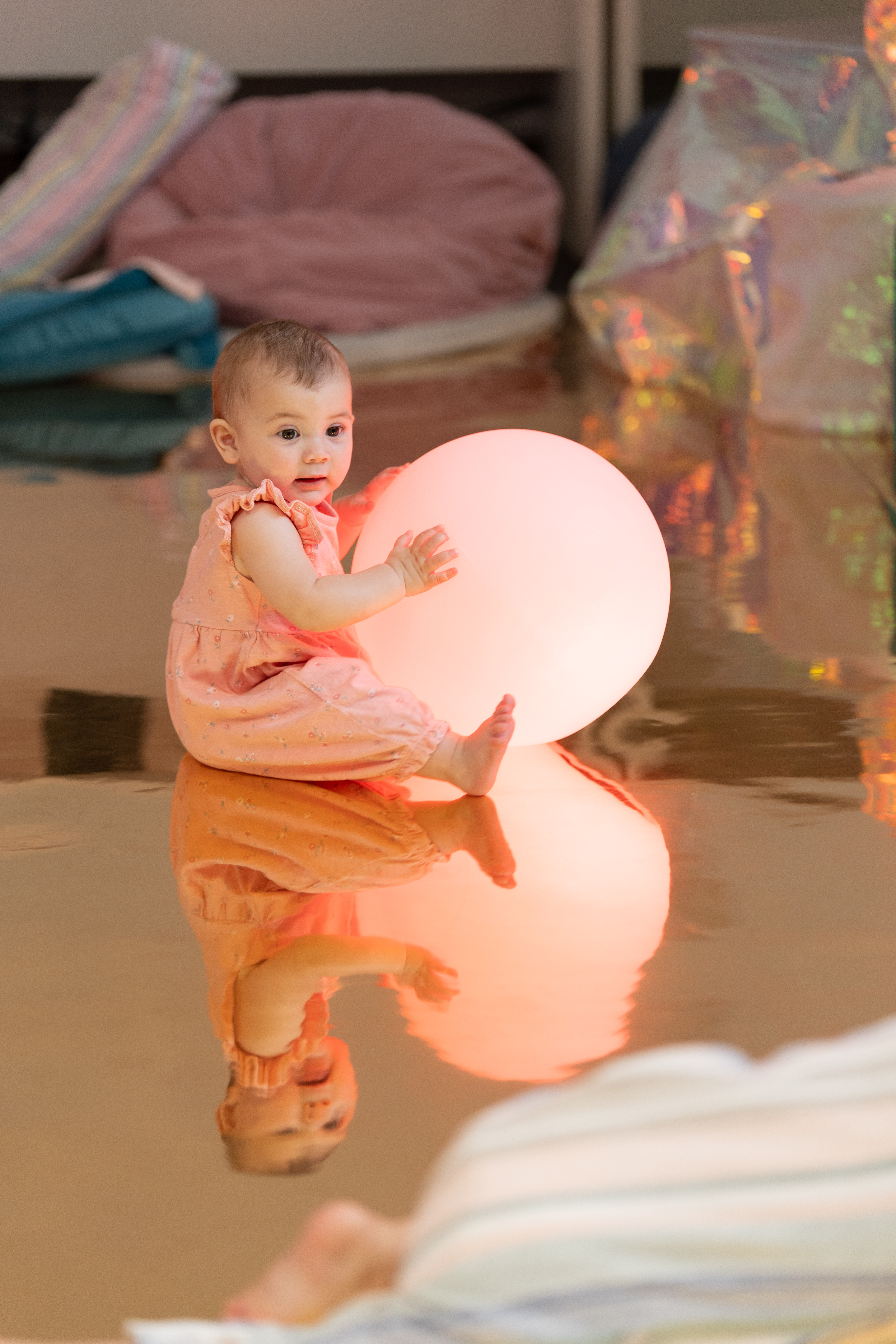 A baby sat on a reflective floor and holding a big bright ball 