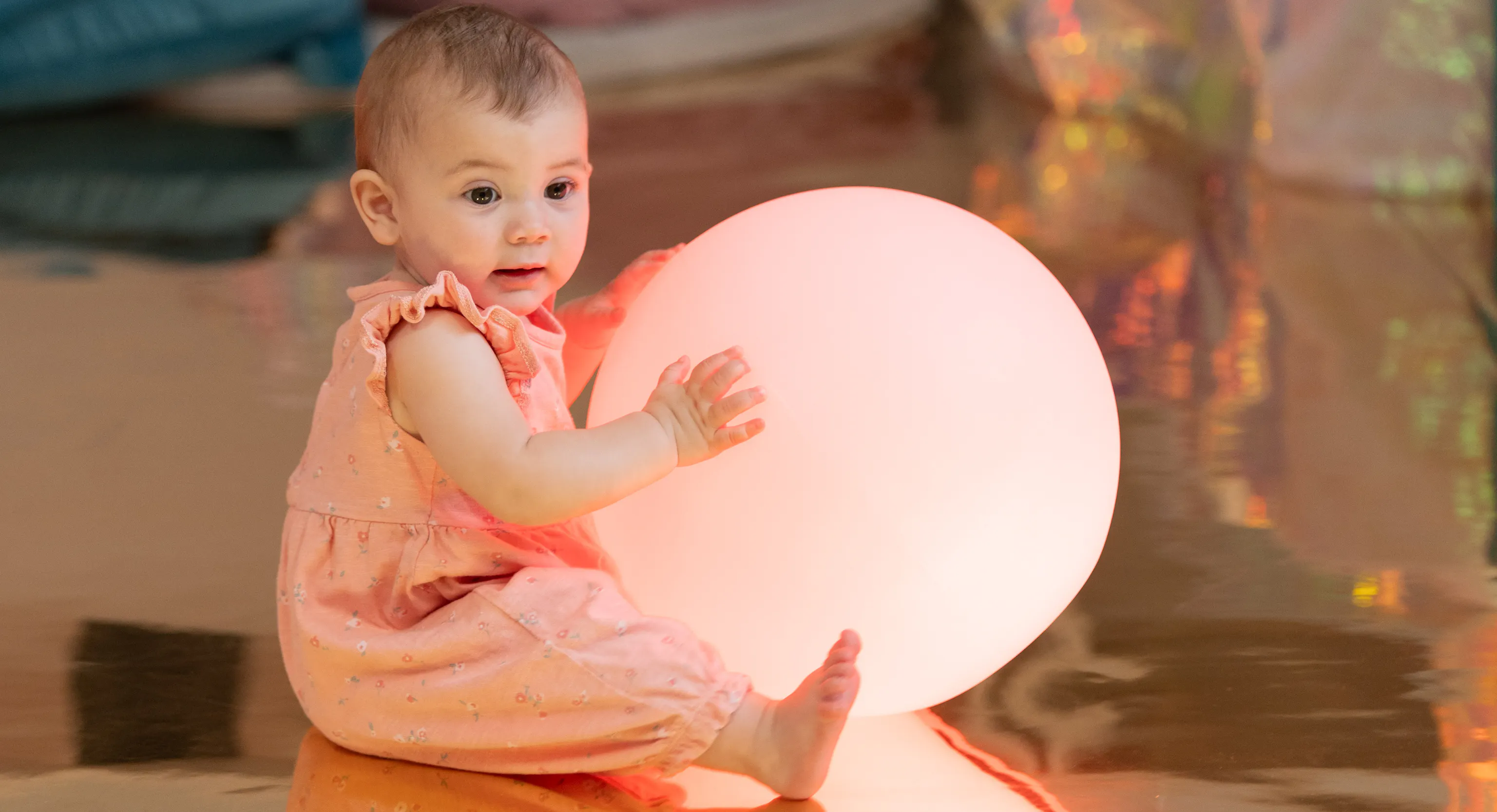 A baby sat on a reflective floor and holding a big bright ball