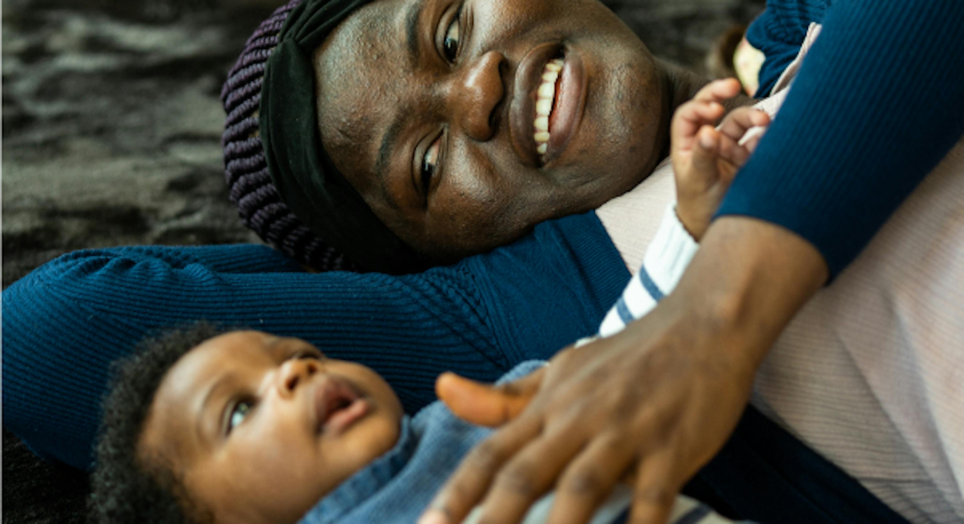 A small baby and their parent/carer are lying on their backs on a black textured mat. The parent/carer is touching the baby's chest who is looking up at the ceiling