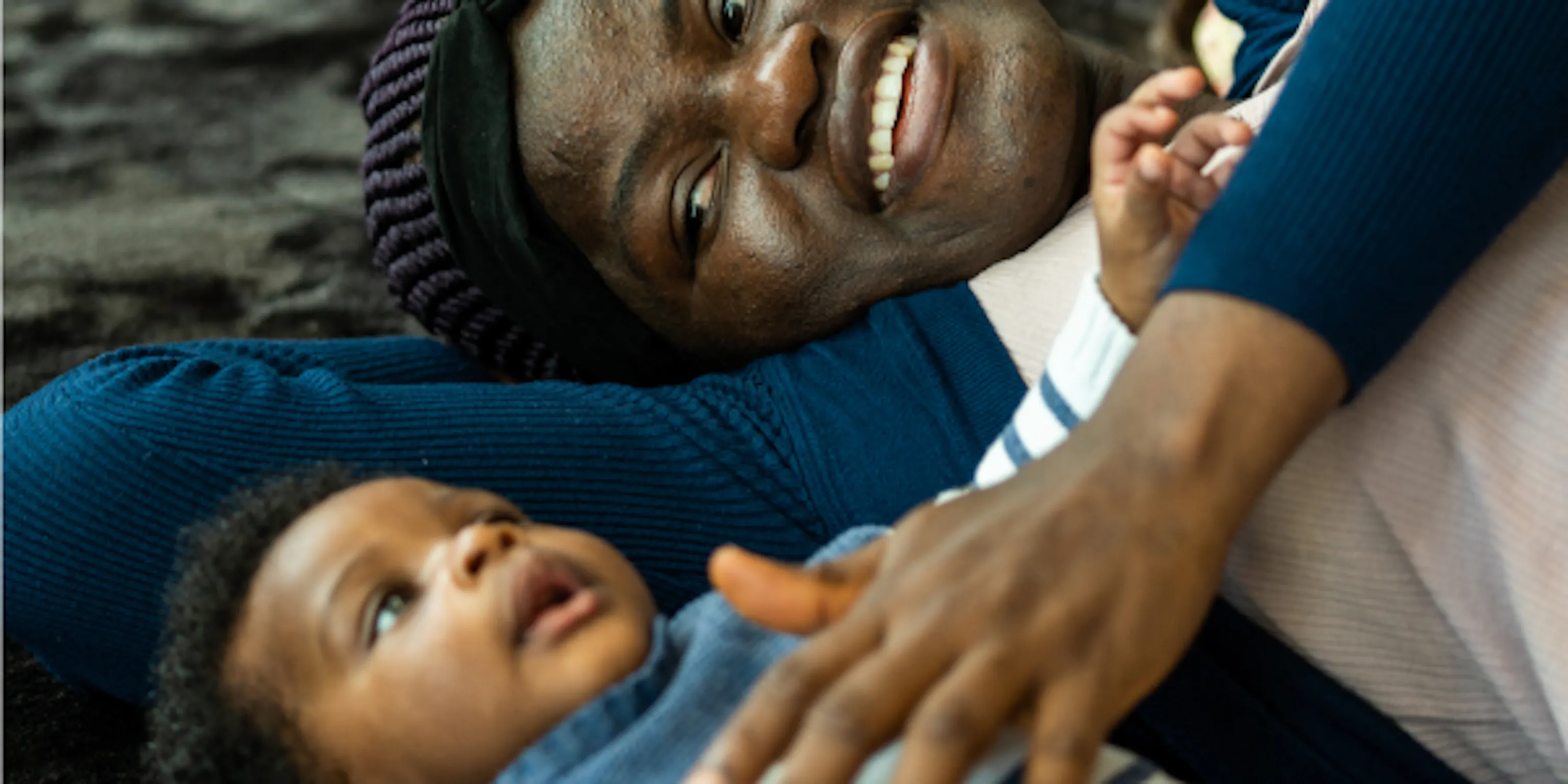 A small baby and their parent/carer are lying on their backs on a black textured mat. The parent/carer is touching the baby's chest who is looking up at the ceiling