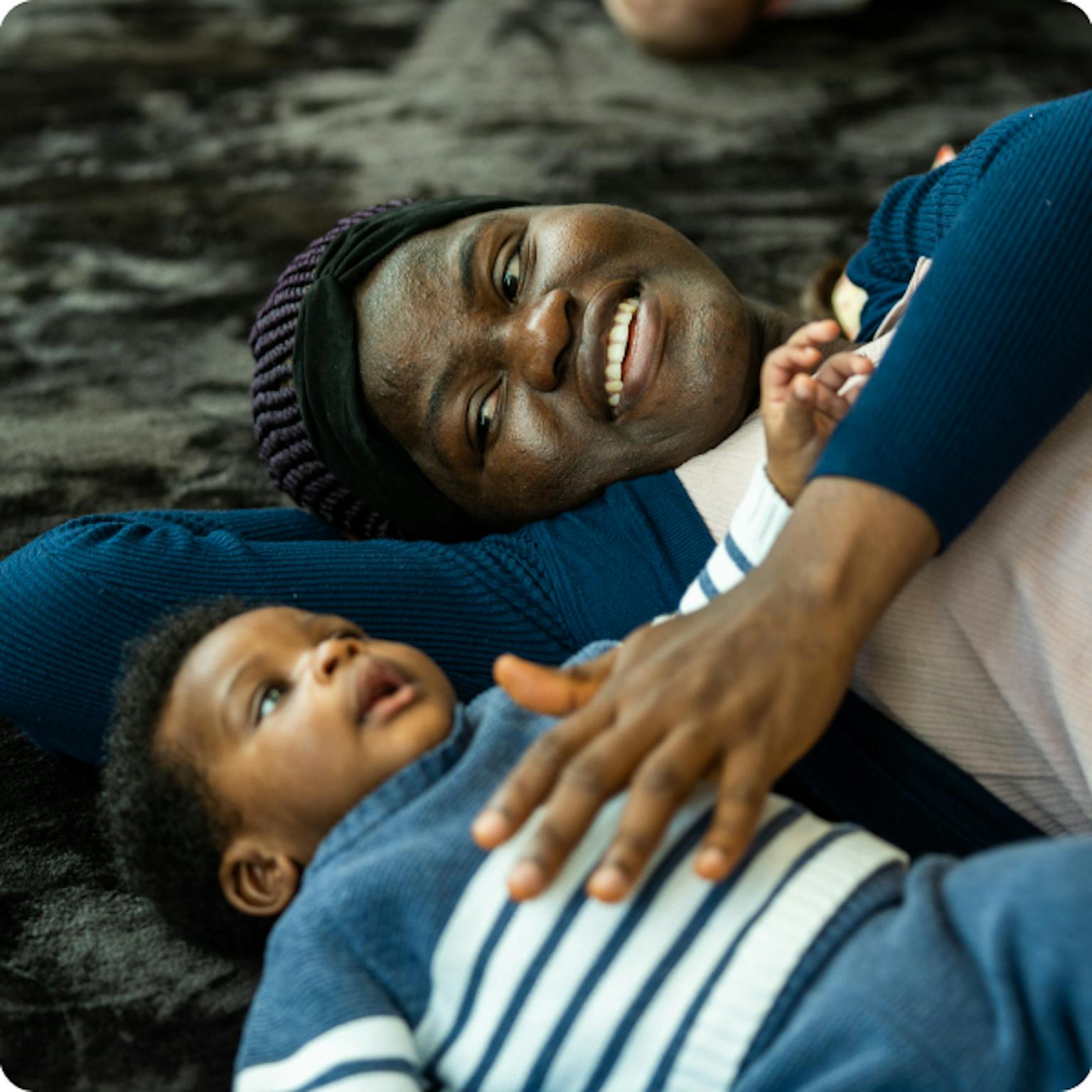 A small baby and their parent/carer are lying on their backs on a black textured mat. The parent/carer is touching the baby's chest who is looking up at the ceiling