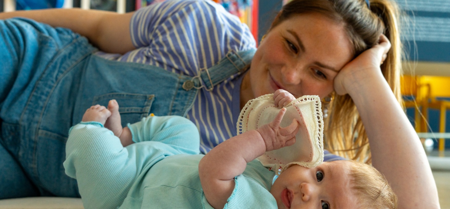 A parent/carer is lying on their side, looking at the baby and smiling. The baby is lying on their back and looking to the camera. They are holding some textured material in their hands. 