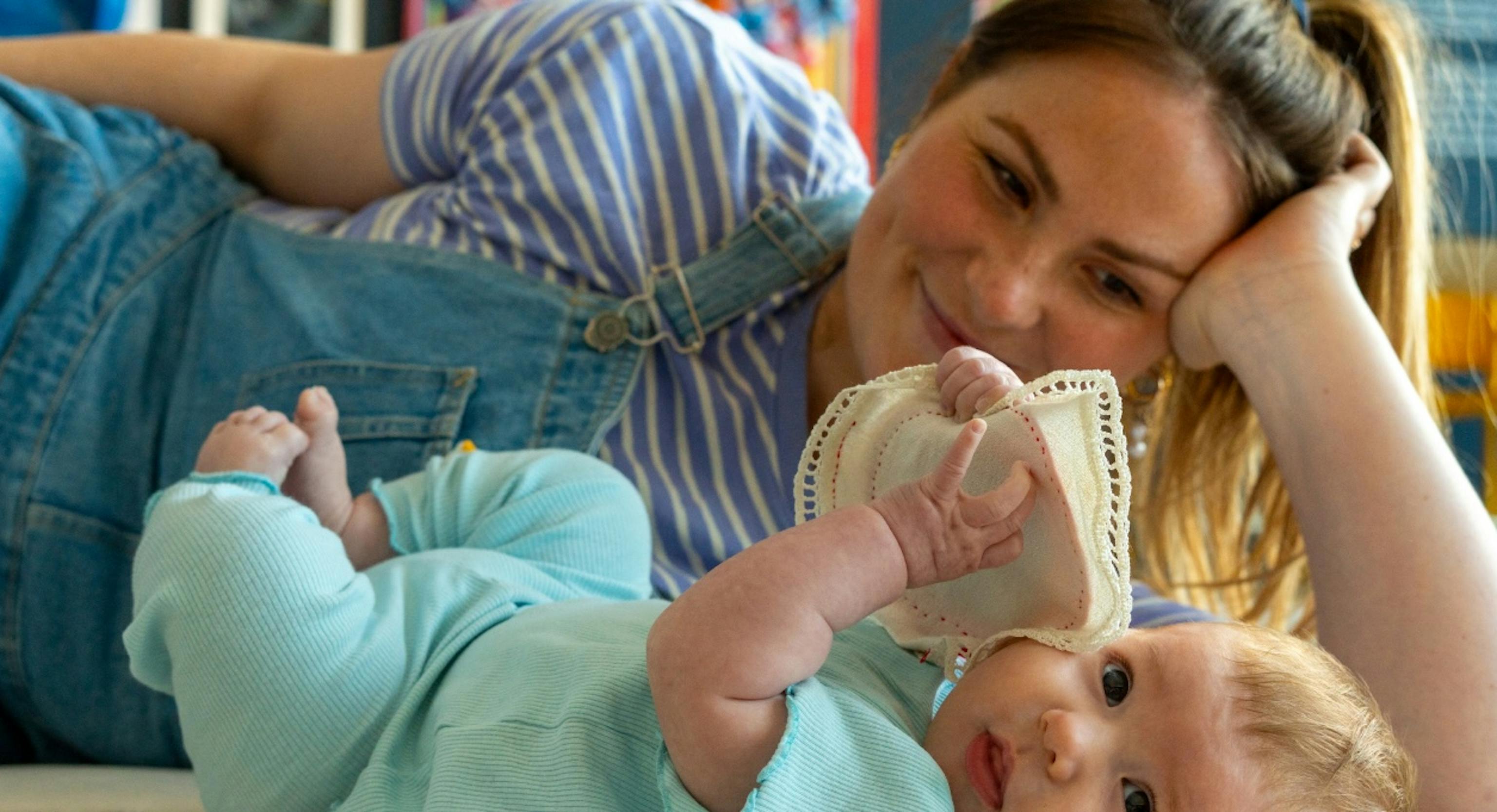 A baby and parent/carer lying on the floor together. The baby is looking towards the camera, holding a piece of material whilst the parent/carer is behind them, looking at the baby and smiling