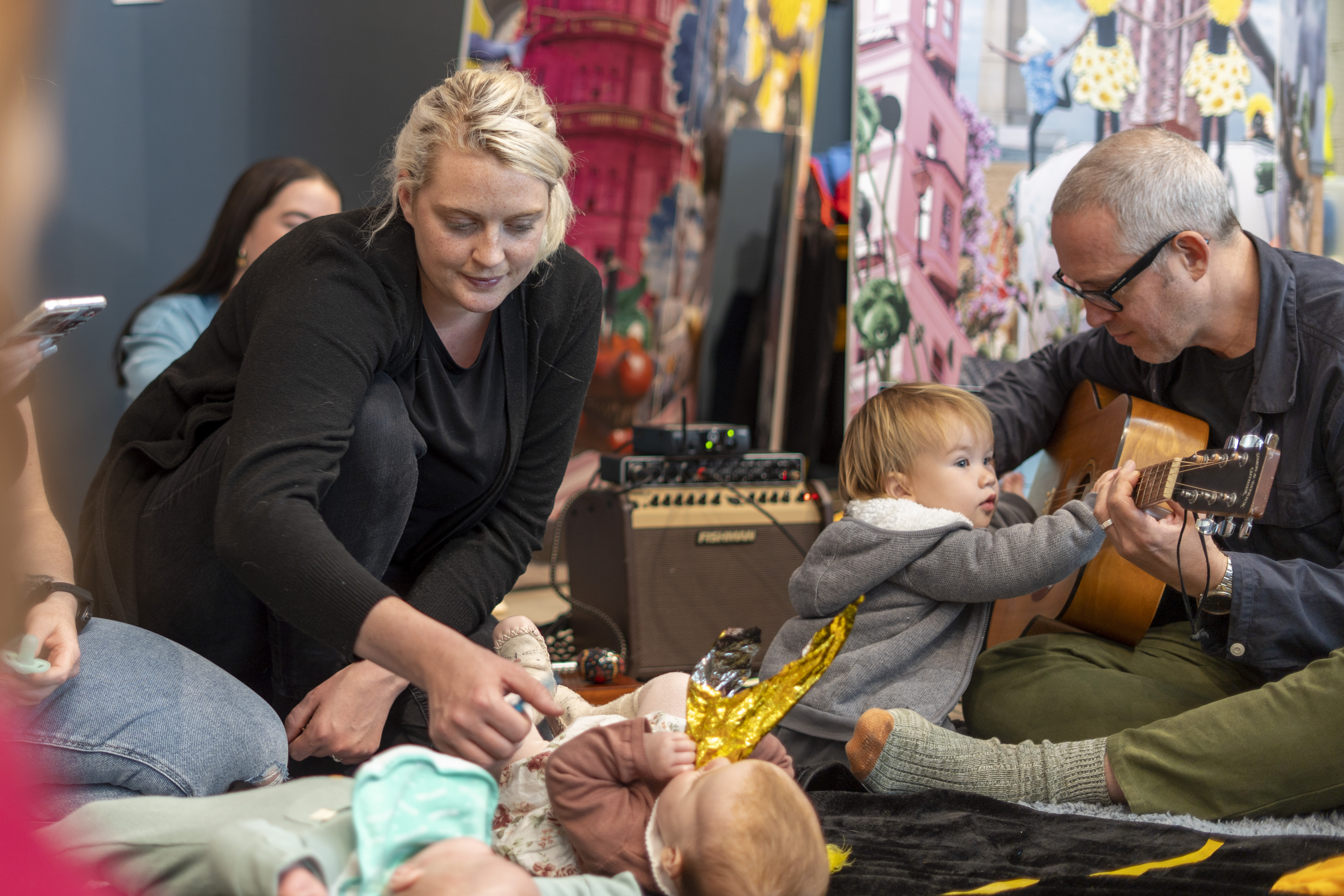 A parent/carer is kneeing and touching a baby that is lying on a textured mate. Next to them, an artist is sitting down playing a guitar and there's baby with them touching the guitar