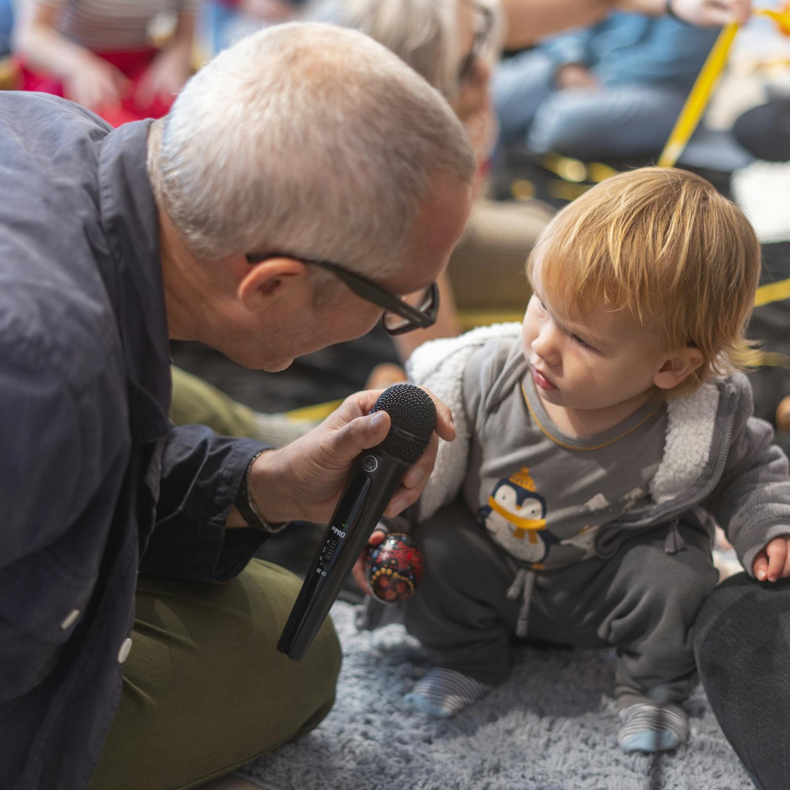 A baby and artist engaging with each other. The artist is holding up a microphone whilst the baby is squatting and looking at the artist