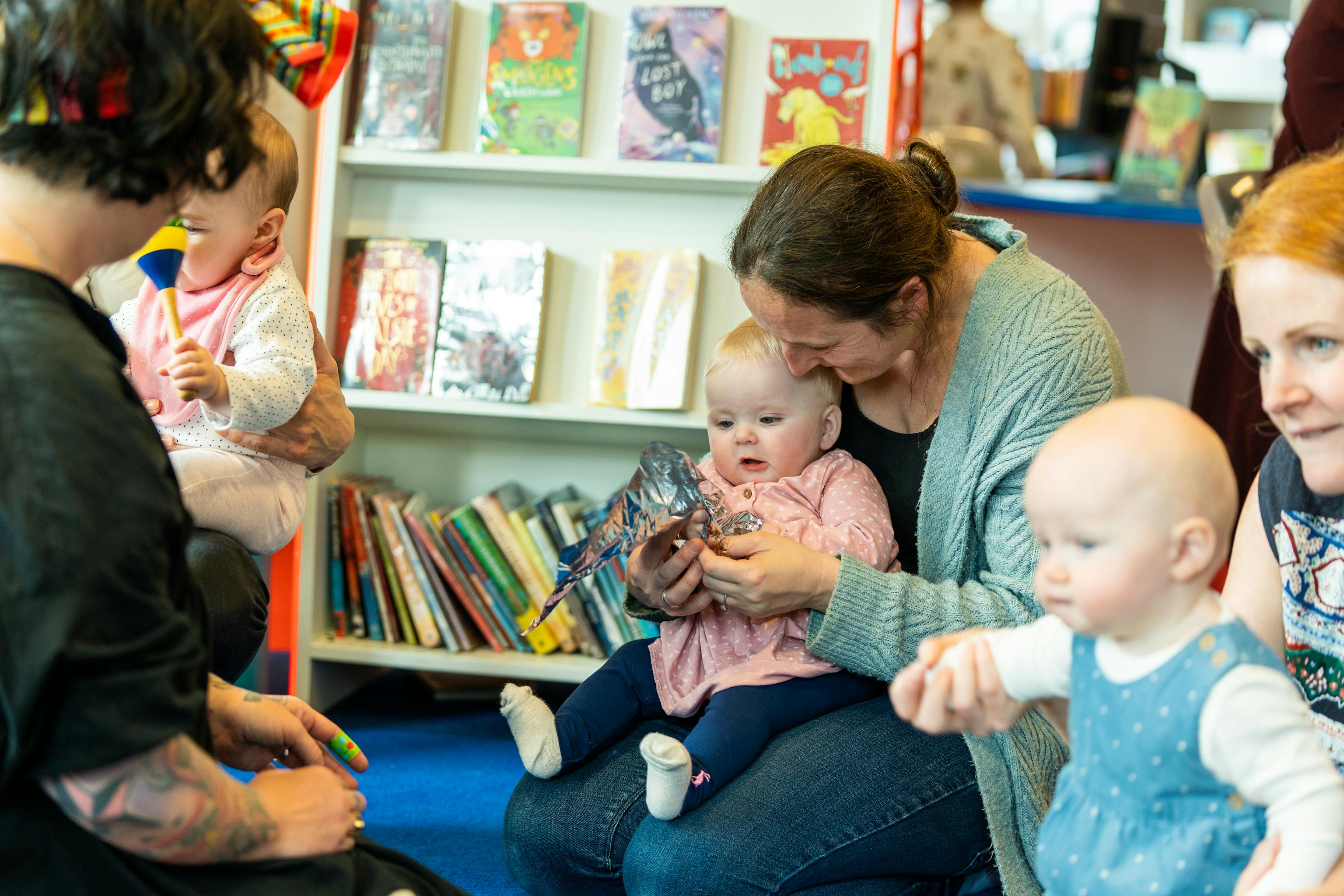 A group of young babies and their parents/carers are sat together during a Baby Playground session in a library