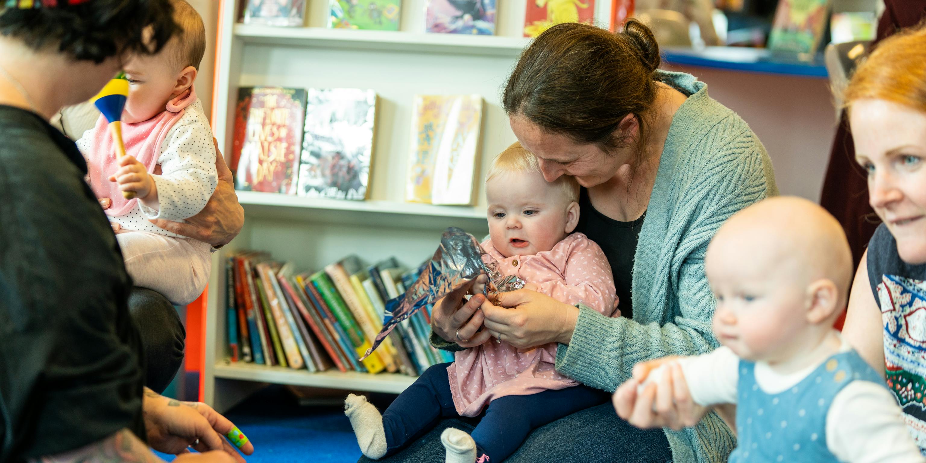 A group of young babies and their parents/carers are sat together during a Baby Playground session in a library