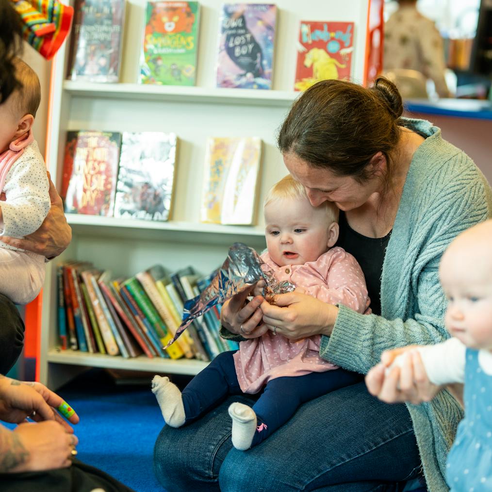 A group of young babies and their parents/carers are sat together during a Baby Playground session in a library
