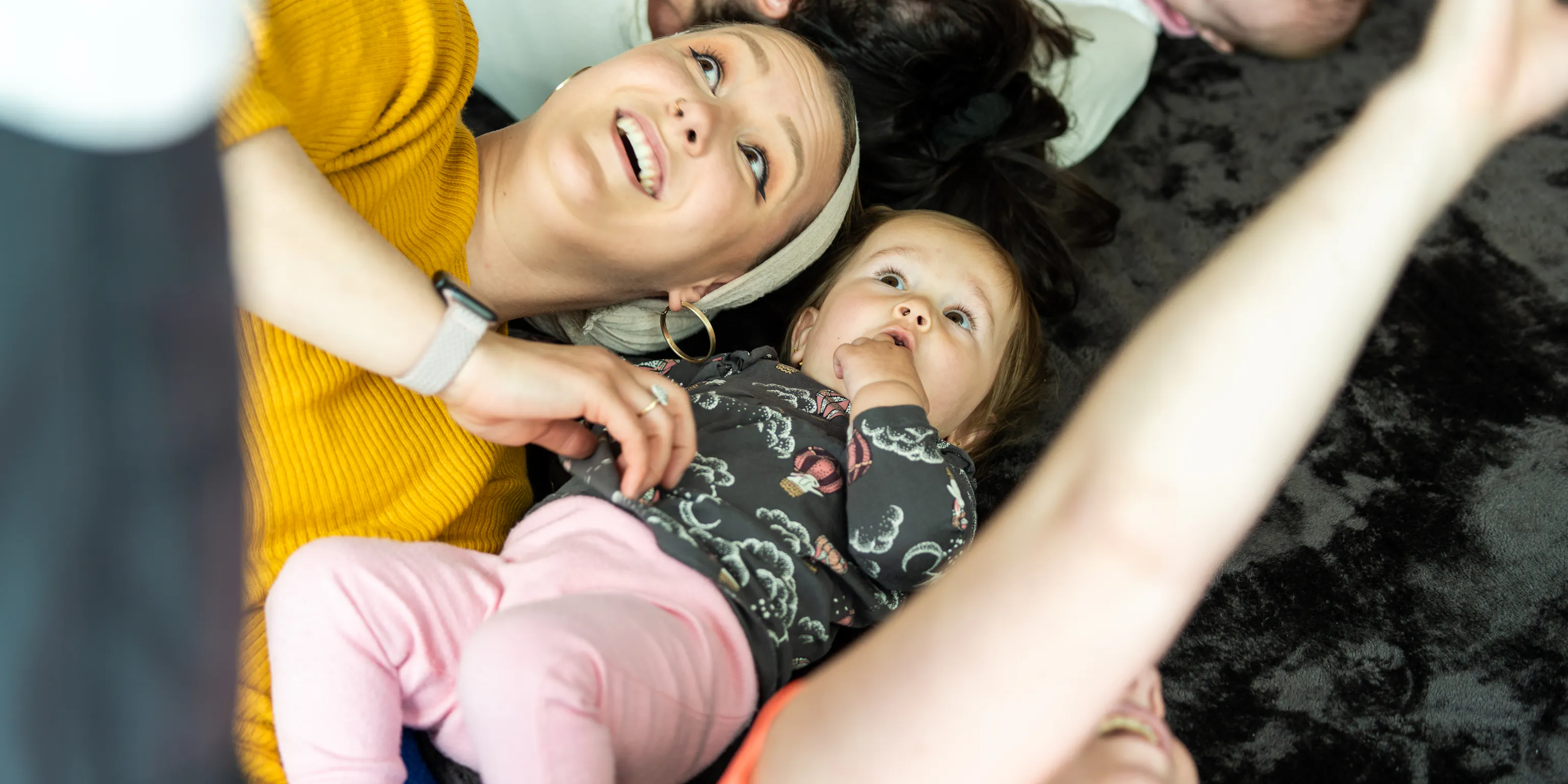 A baby and their parent/carer are lying on a black textured mat together and looking at the ceiling and smiling. Next to them is someone with their hands up reaching for something out of the camera's focus