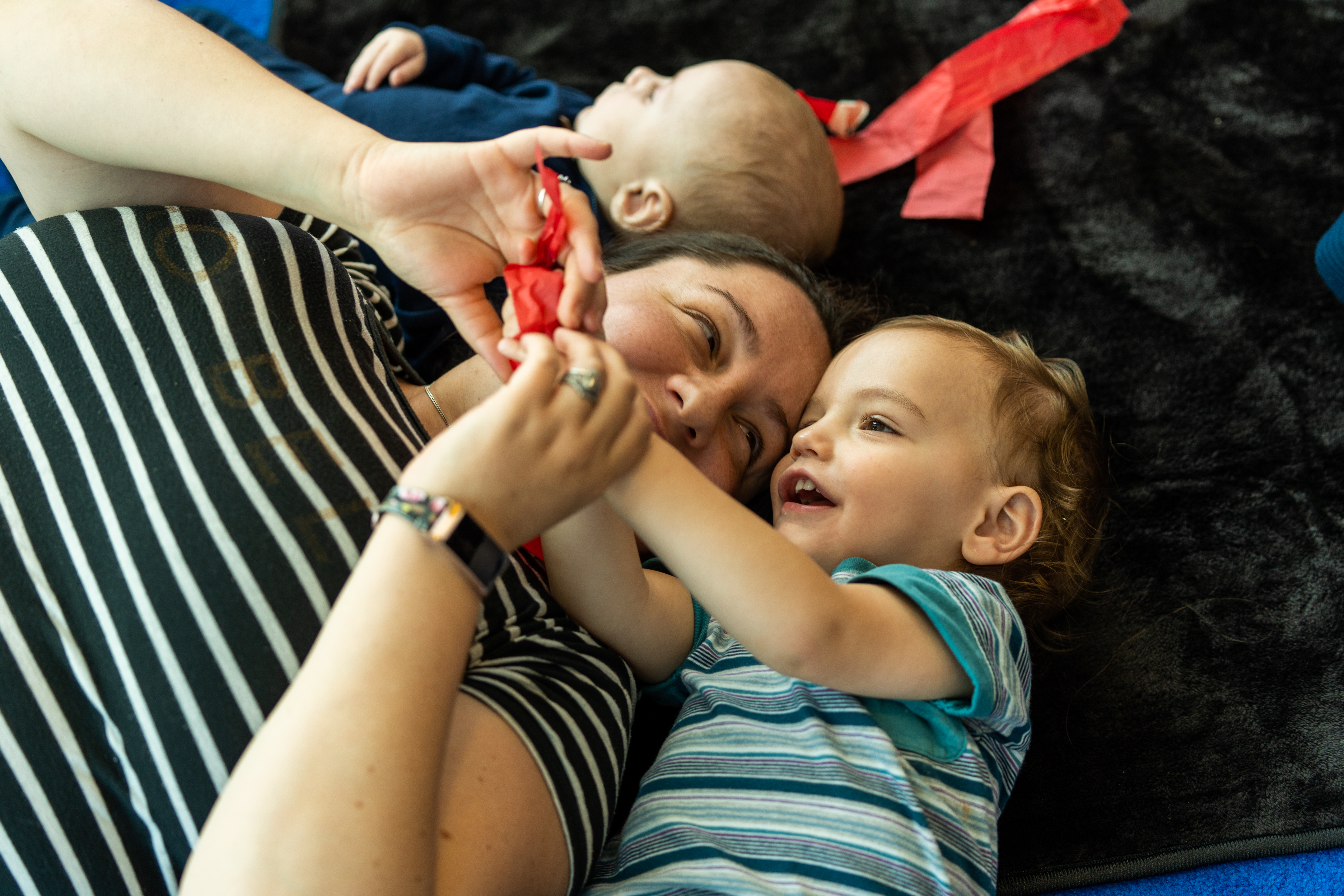 A parent/carer is lying on a textured black mat next to a young child. They are both holding a red piece of tissue paper and smiling.