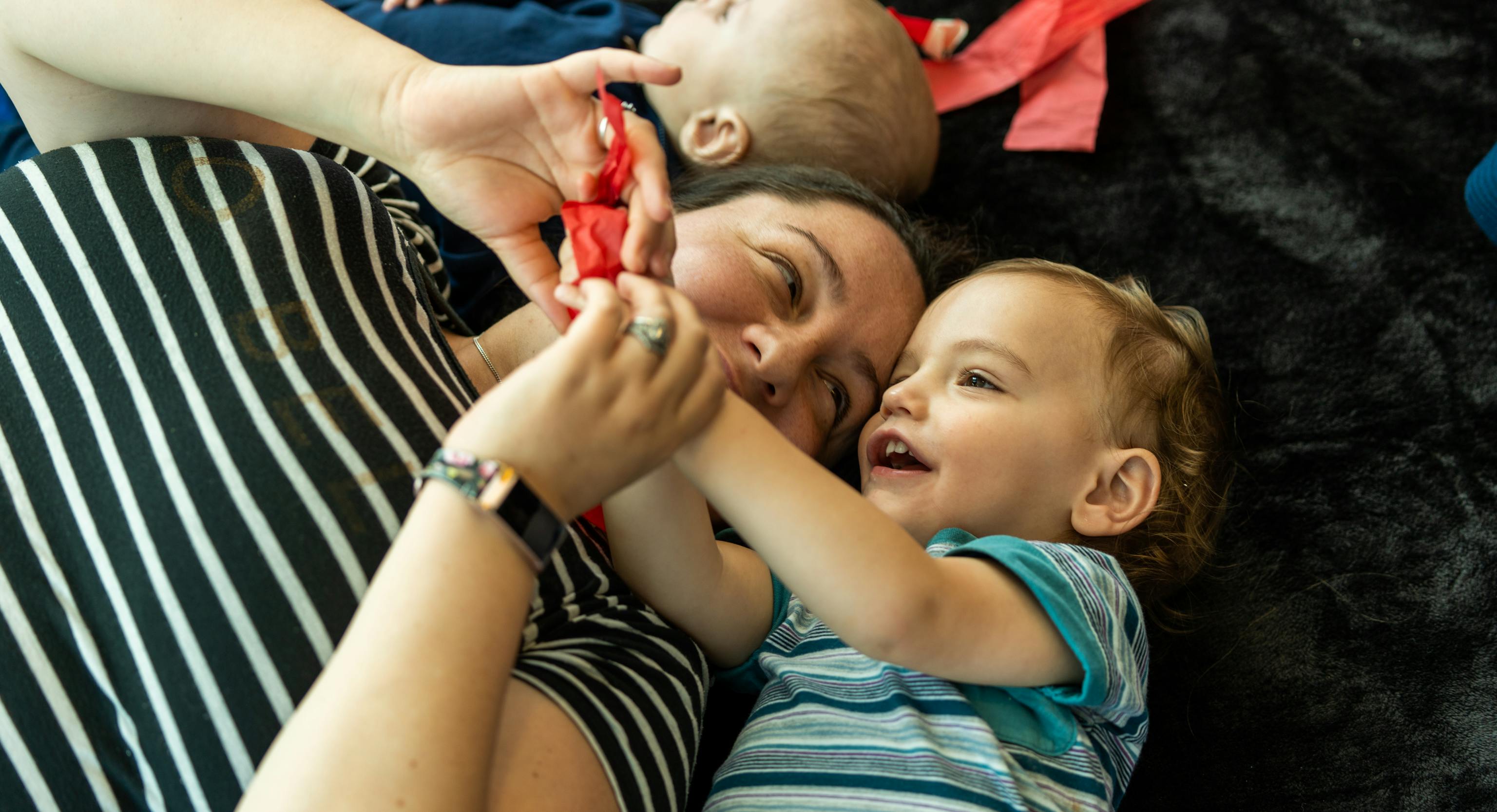 A parent/carer is lying on a textured black mat next to a young child. They are both holding a red piece of tissue paper and smiling.