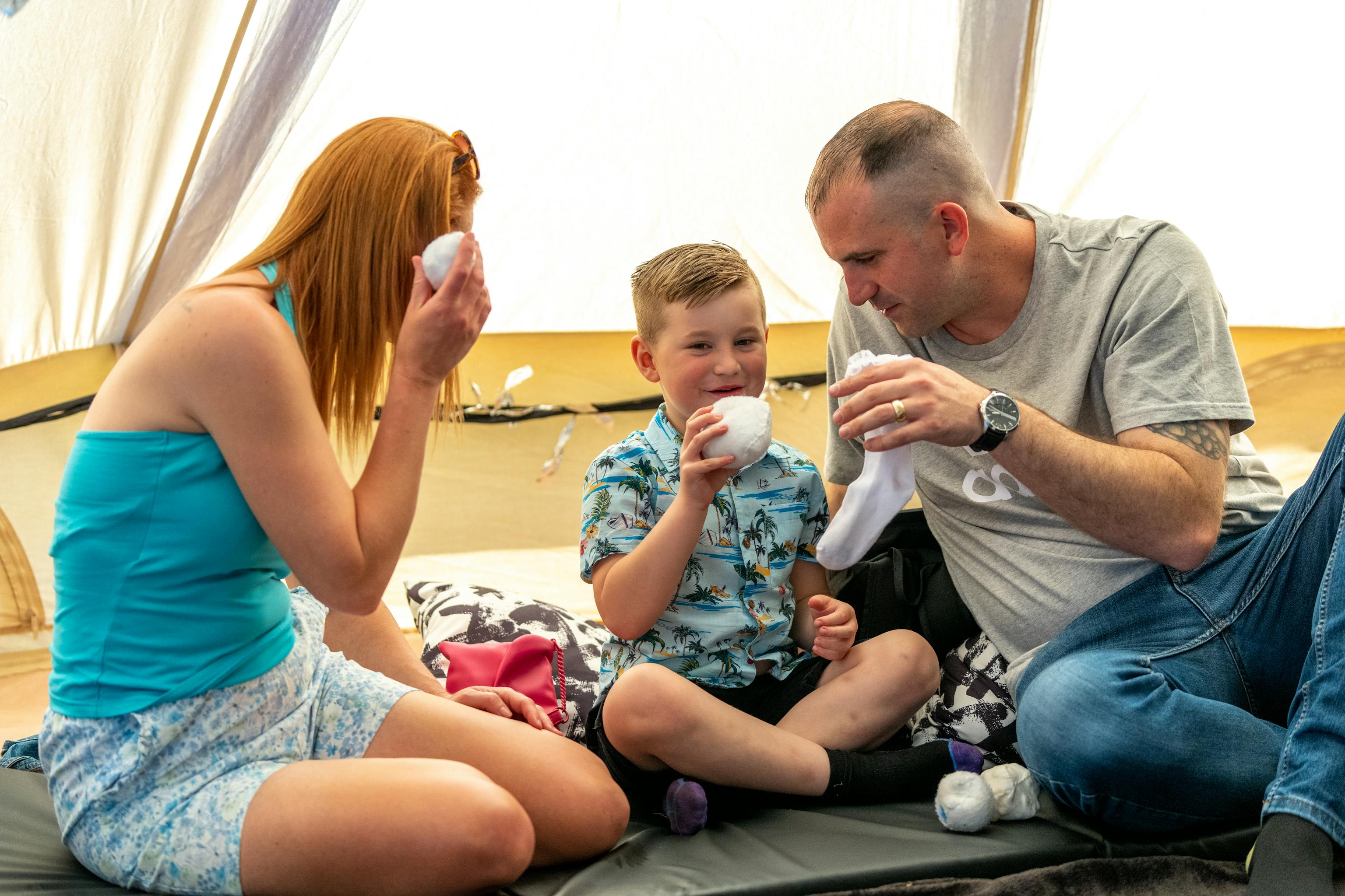 Two parents/carers are sat with a young child. They are all holding small white balls and playing with them inside a tipi tent at a festival