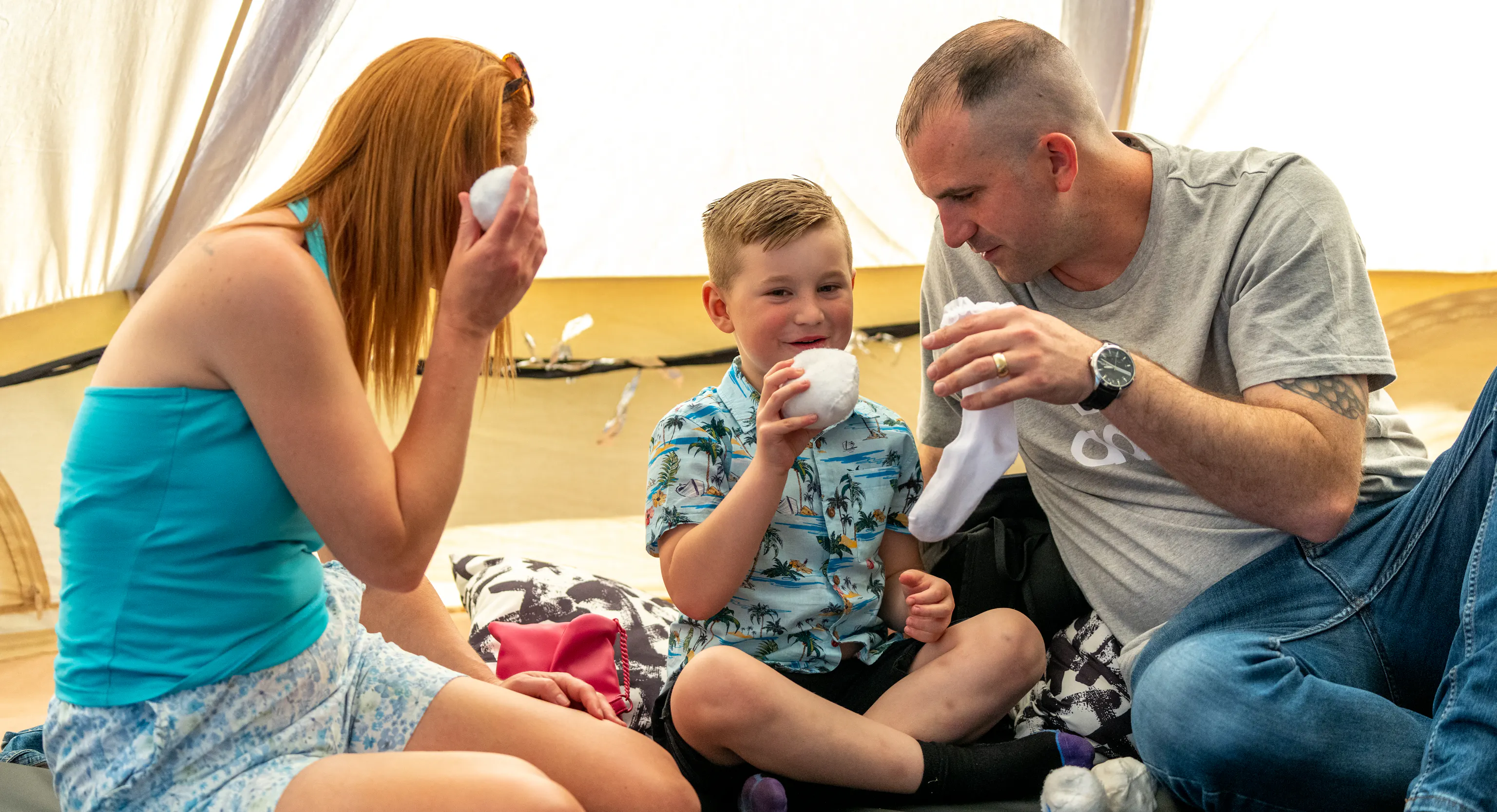 A family sat together on the floor. The parent/carer to the left is looking at the young child in the middle with a white ball in their hand. The other parent/carer is looking at the child with a sock in their hand. The young child in the middle is smiling and holding a white sock/ball